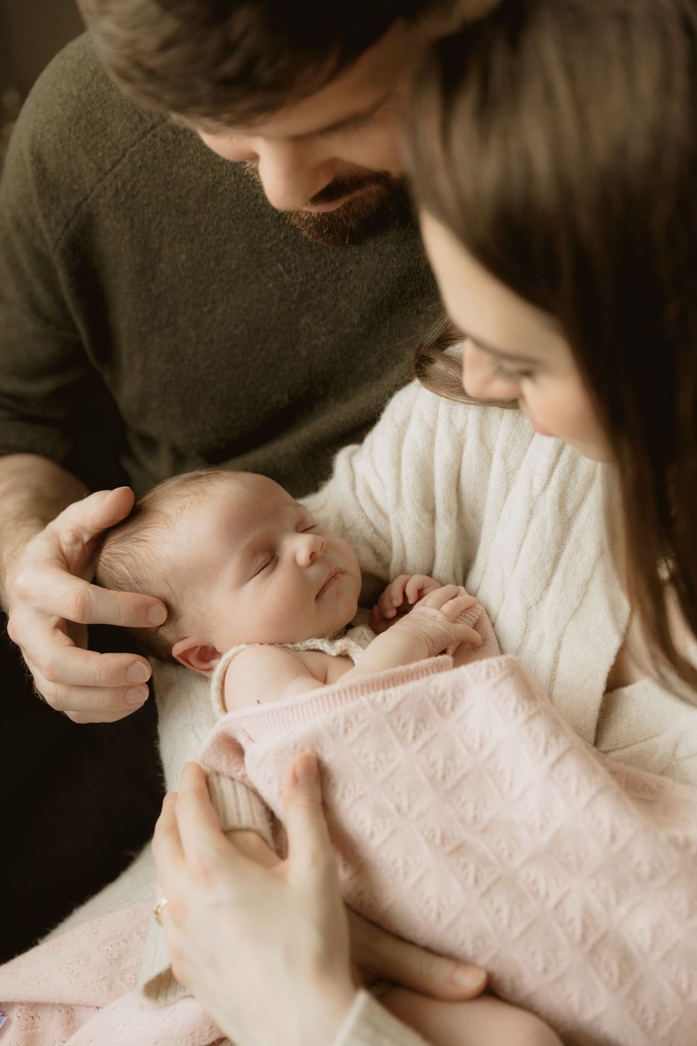 Parents in cozy sweaters gaze lovingly at their sleeping newborn baby wrapped in a soft pink blanket.