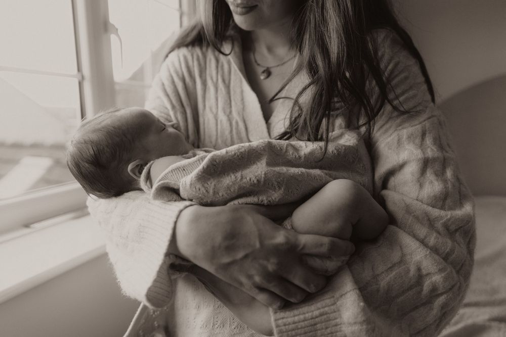 A person sits by a window holding a swaddled baby in a nurturing black and white photograph.