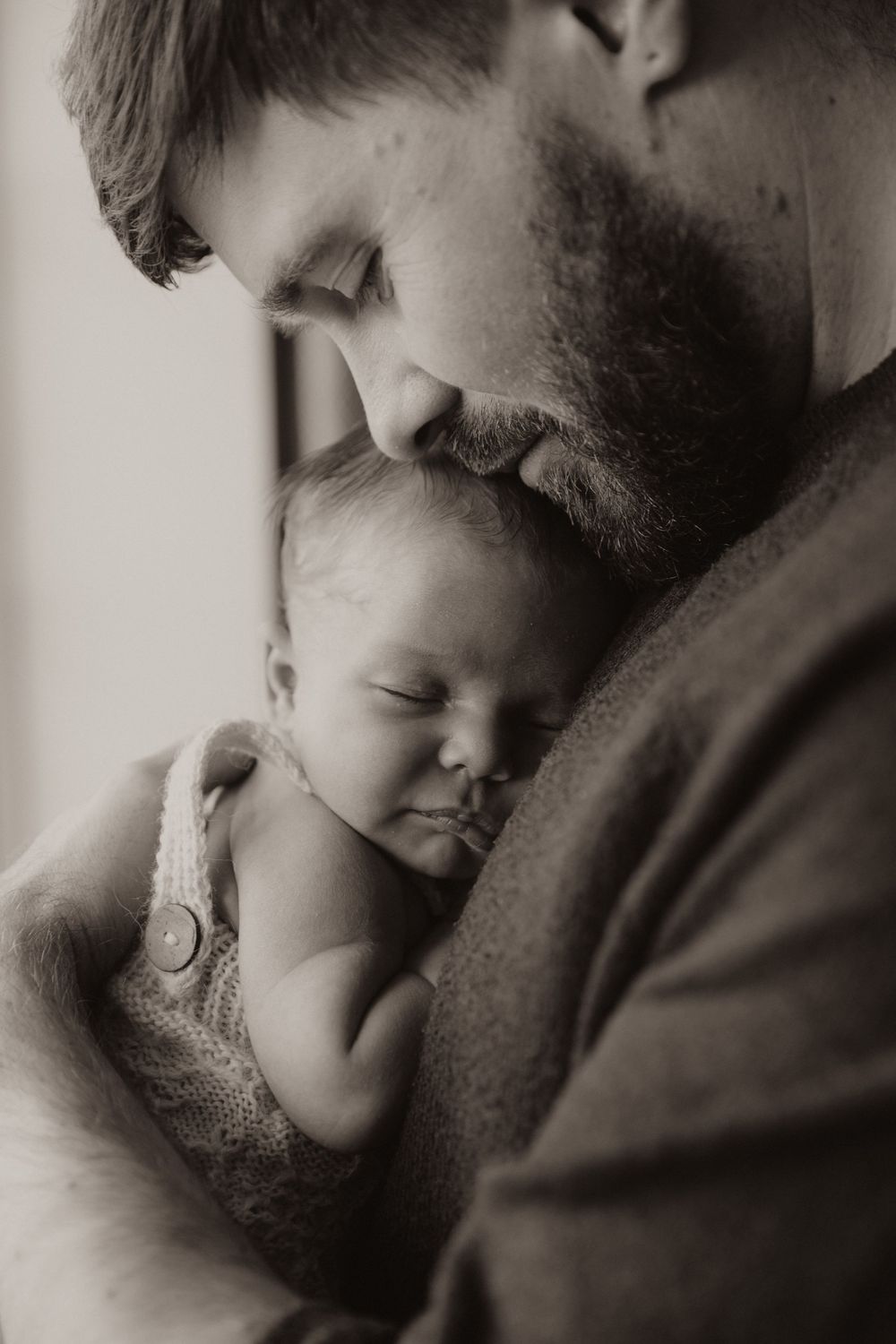 A tender black and white photograph captures a bearded parent gently cradling a sleeping newborn against their chest.