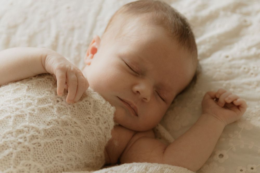 Peaceful sleeping baby lying on white lace blanket with arms outstretched in natural light.