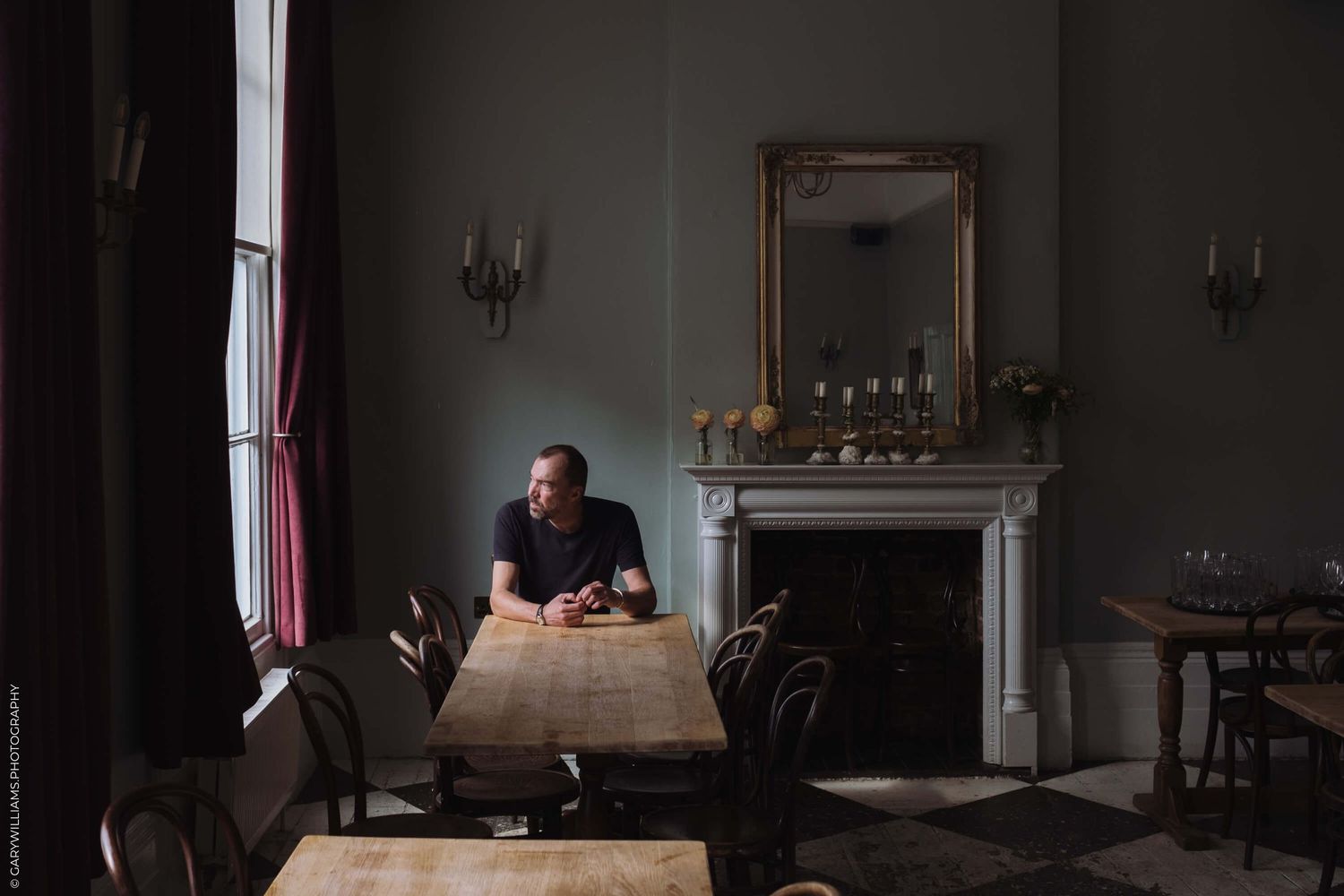 A person sits alone at a wooden table in a moody, dimly lit vintage dining room with ornate fireplace and mirror.