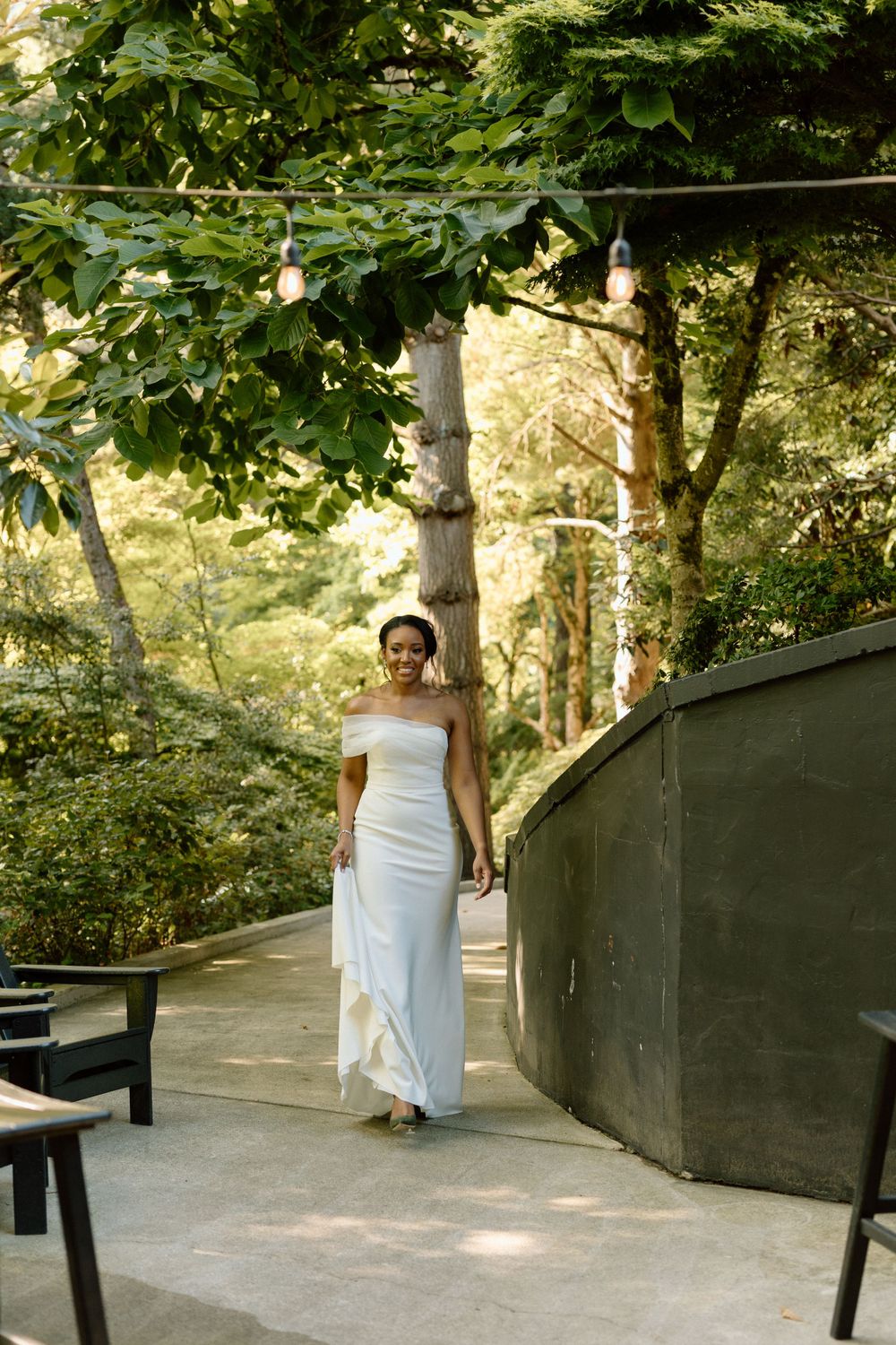 An elegant off-shoulder white wedding dress is showcased on a winding garden path surrounded by lush greenery.