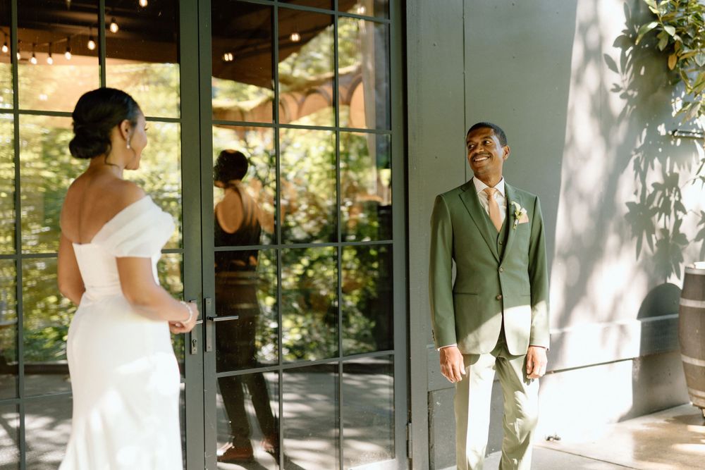 A couple in wedding attire shares a tender moment in front of a sunlit windowed wall with greenery backdrop.