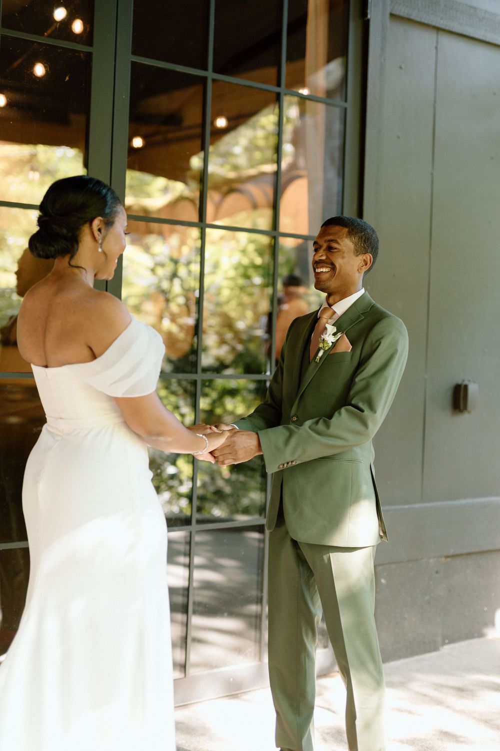 Two people share a joyful moment during a wedding, with the bride in a white off-shoulder dress and groom in a sage green suit.