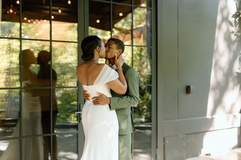 A couple shares an intimate moment in their wedding attire while dancing in front of a sunlit glass window.