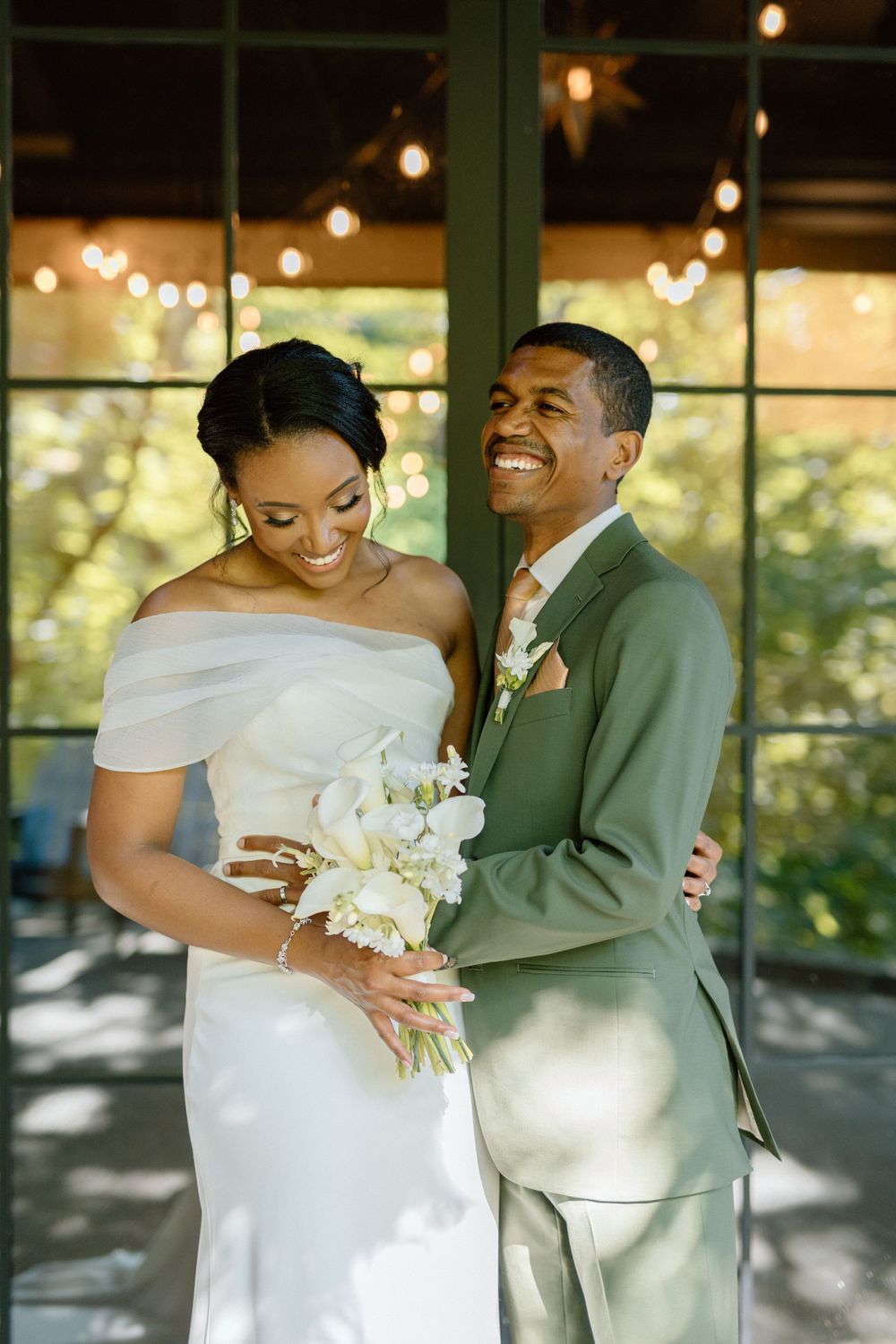 Elegant wedding couple shares a joyful moment together against string lights and greenery backdrop.