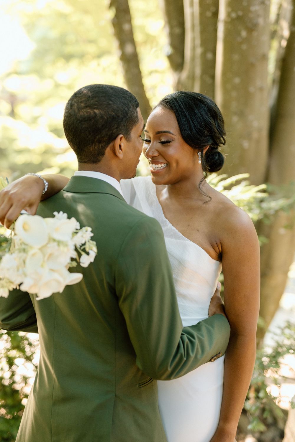 A smiling bride in a white dress shares a romantic moment with her groom in a green suit in a sunlit forest setting.