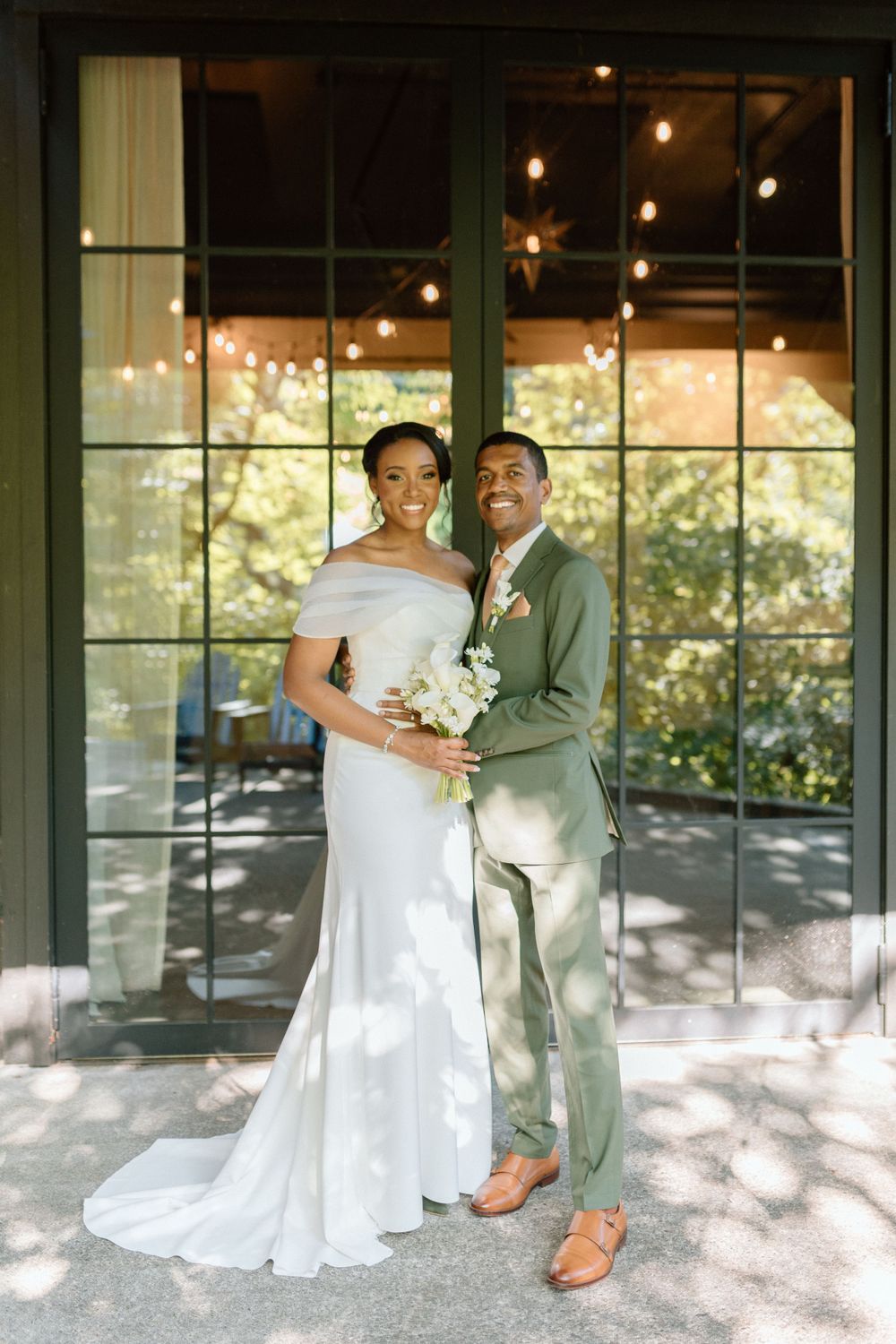 A couple poses in formal wedding attire in front of large windows with string lights and greenery in the background.