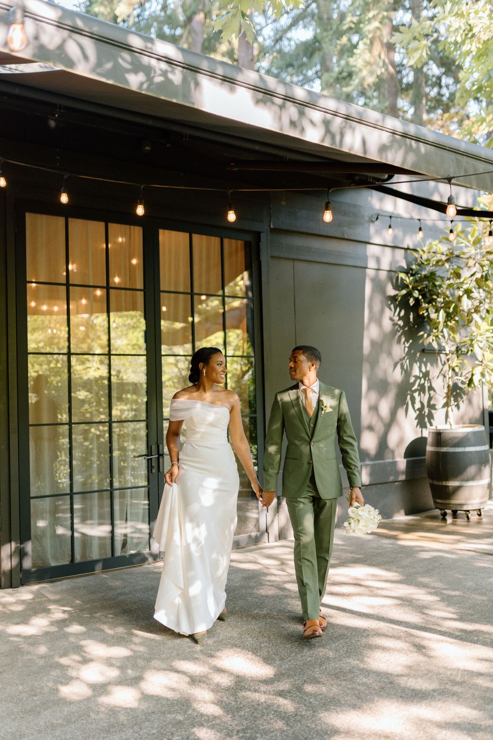 A couple in wedding attire walks together outside a modern building with string lights and glass windows in dappled sunlight.