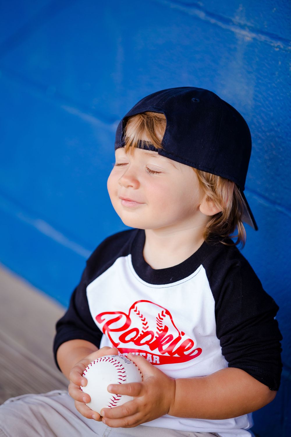 Child in baseball attire holds baseball while wearing Rookie gear and baseball cap against blue backdrop.