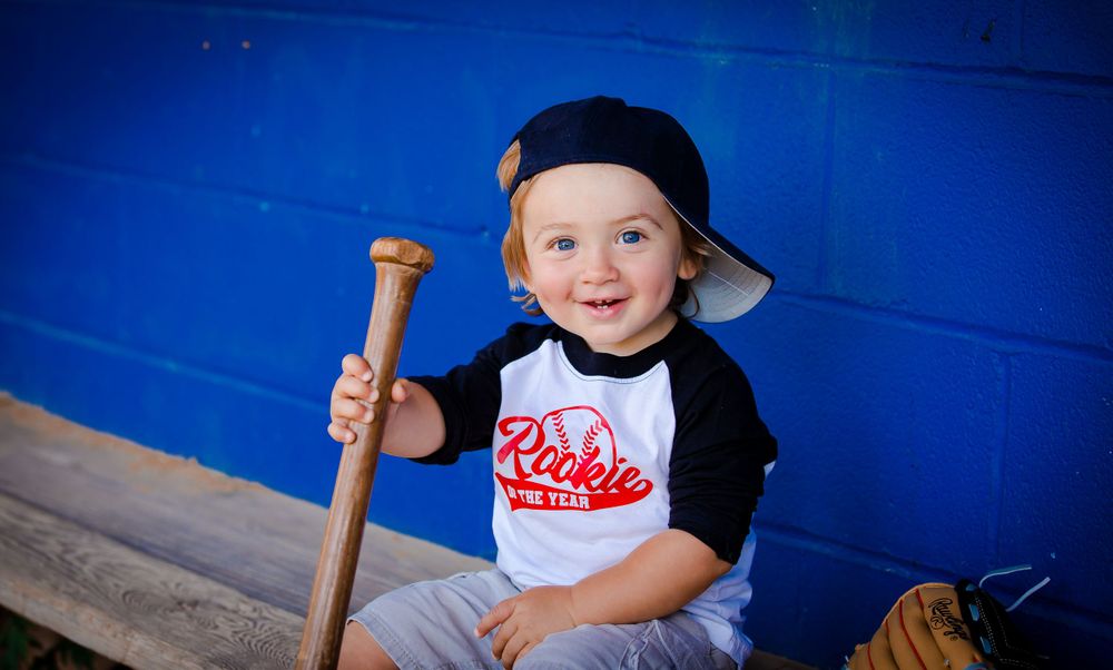 Young child in baseball attire sits in dugout holding wooden bat against bright blue background.