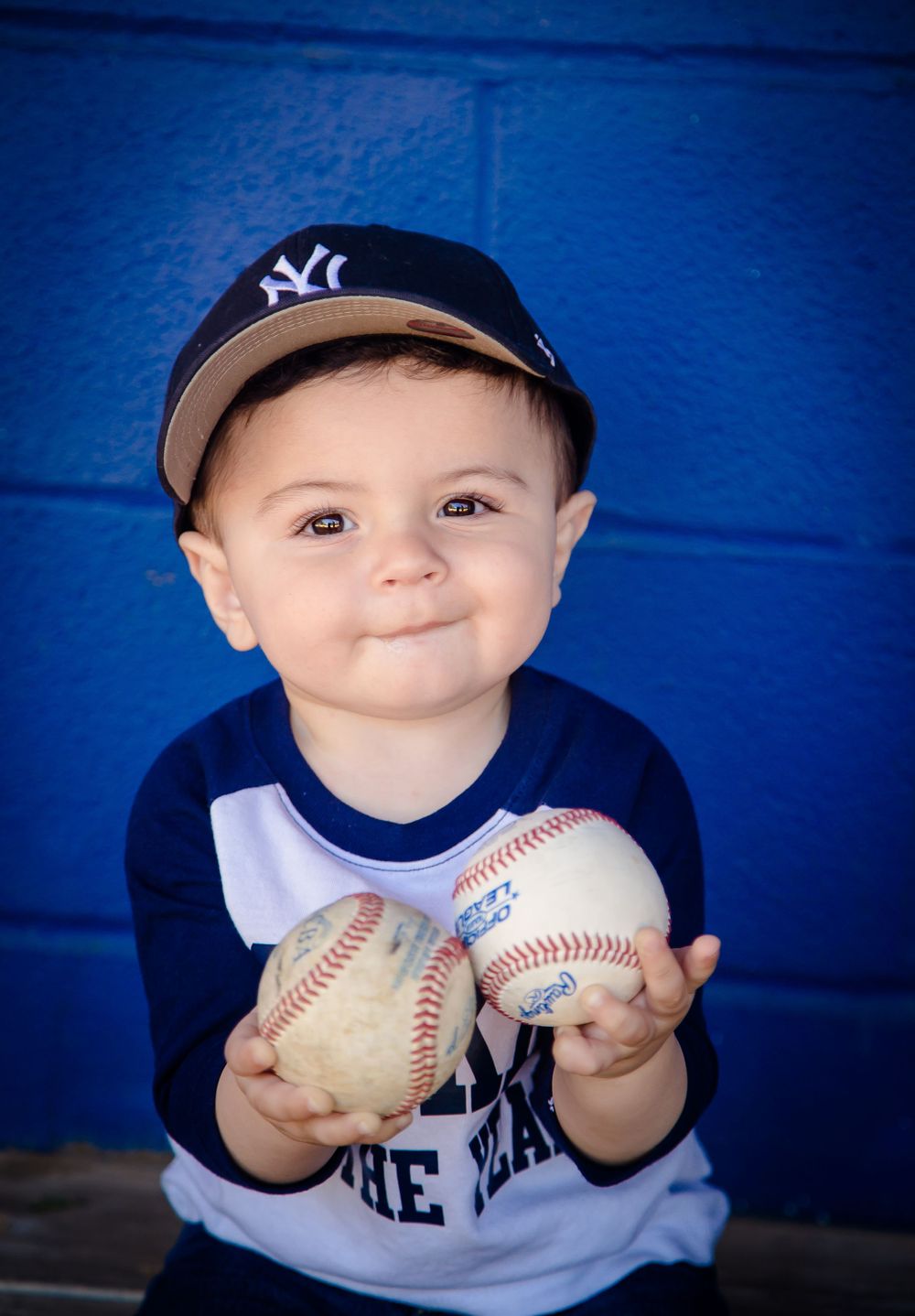A young baseball fan wearing a Yankees cap and team shirt holds two baseballs against a bright blue wall background.