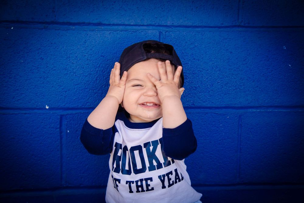 Small child in baseball shirt covering eyes while laughing against bright blue wall background.