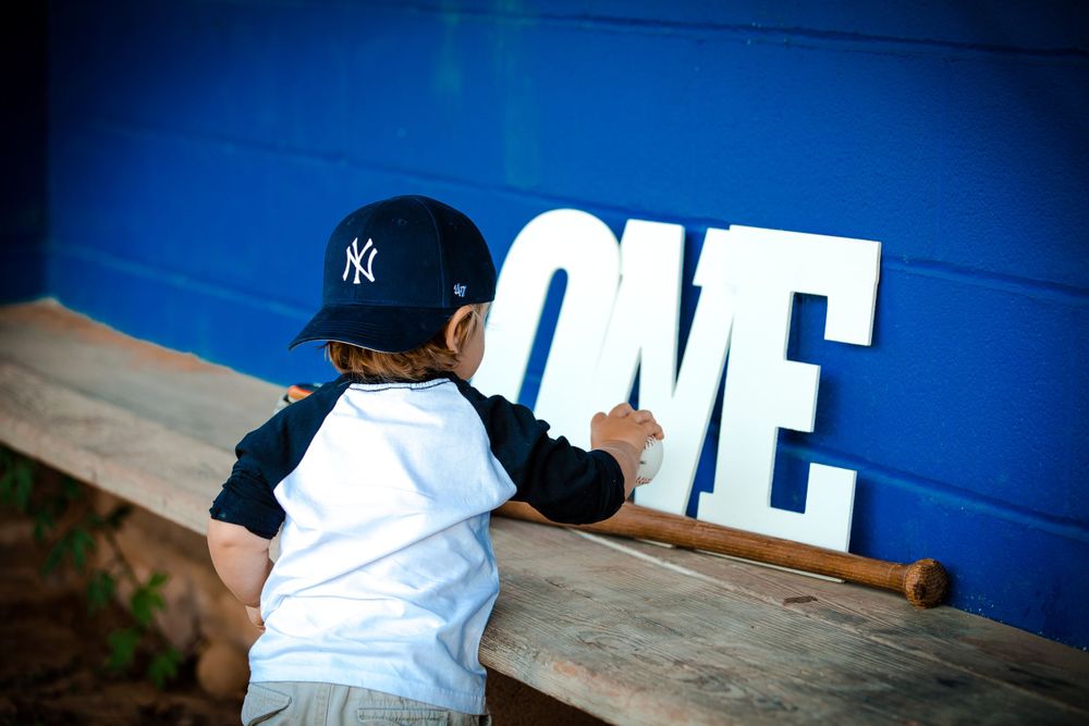 A young baserunner wearing a New York Yankees cap poses next to large white letters spelling 'ONE' against a blue wall.