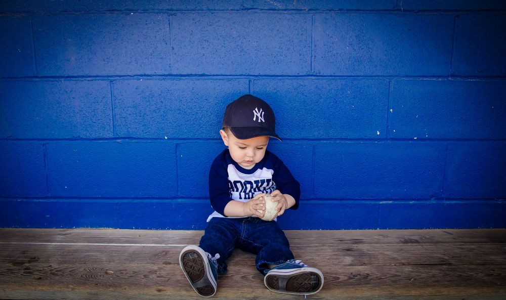 A small fan wearing a Yankees cap and baseball uniform sits against a bright blue wall.