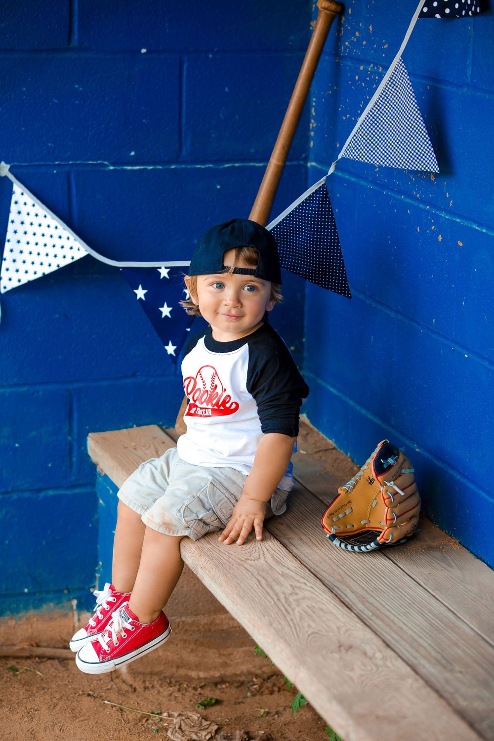 A young baseball player sits on a dugout bench with decorative bunting and a glove nearby against a bright blue wall.