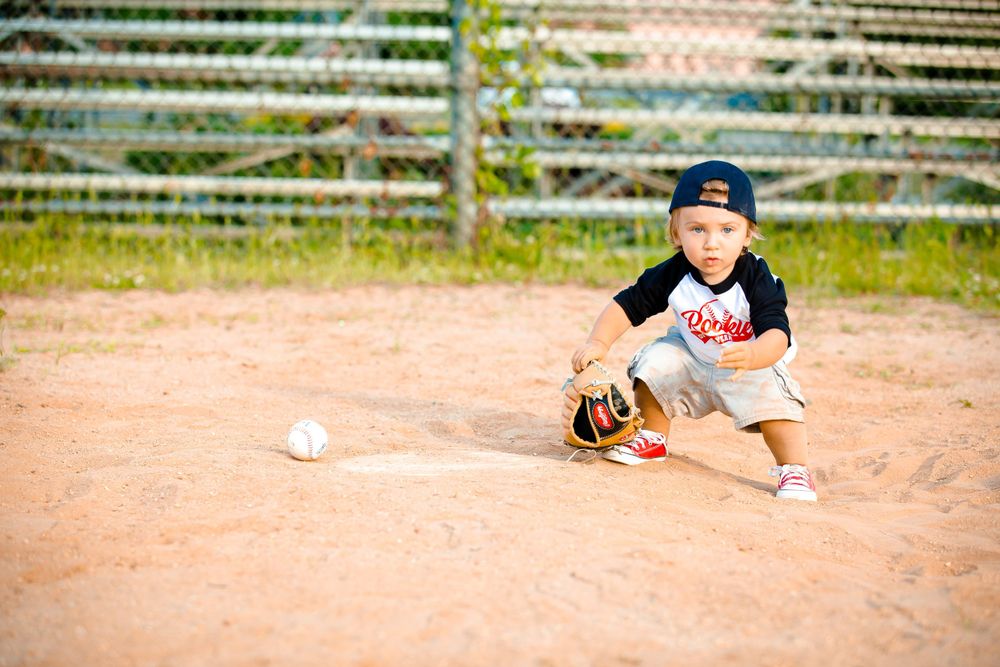 A small baseball player crouches in ready position on a dirt field with a ball nearby and wooden fencing in background.
