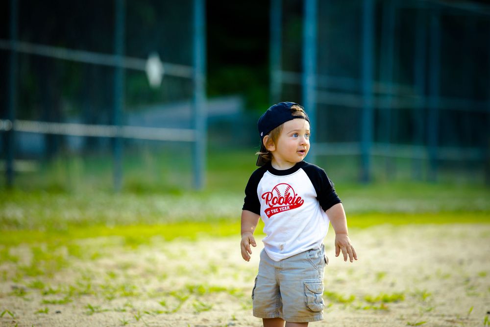 A young child in a baseball style shirt and cap looks up at the sky while standing on a baseball field.