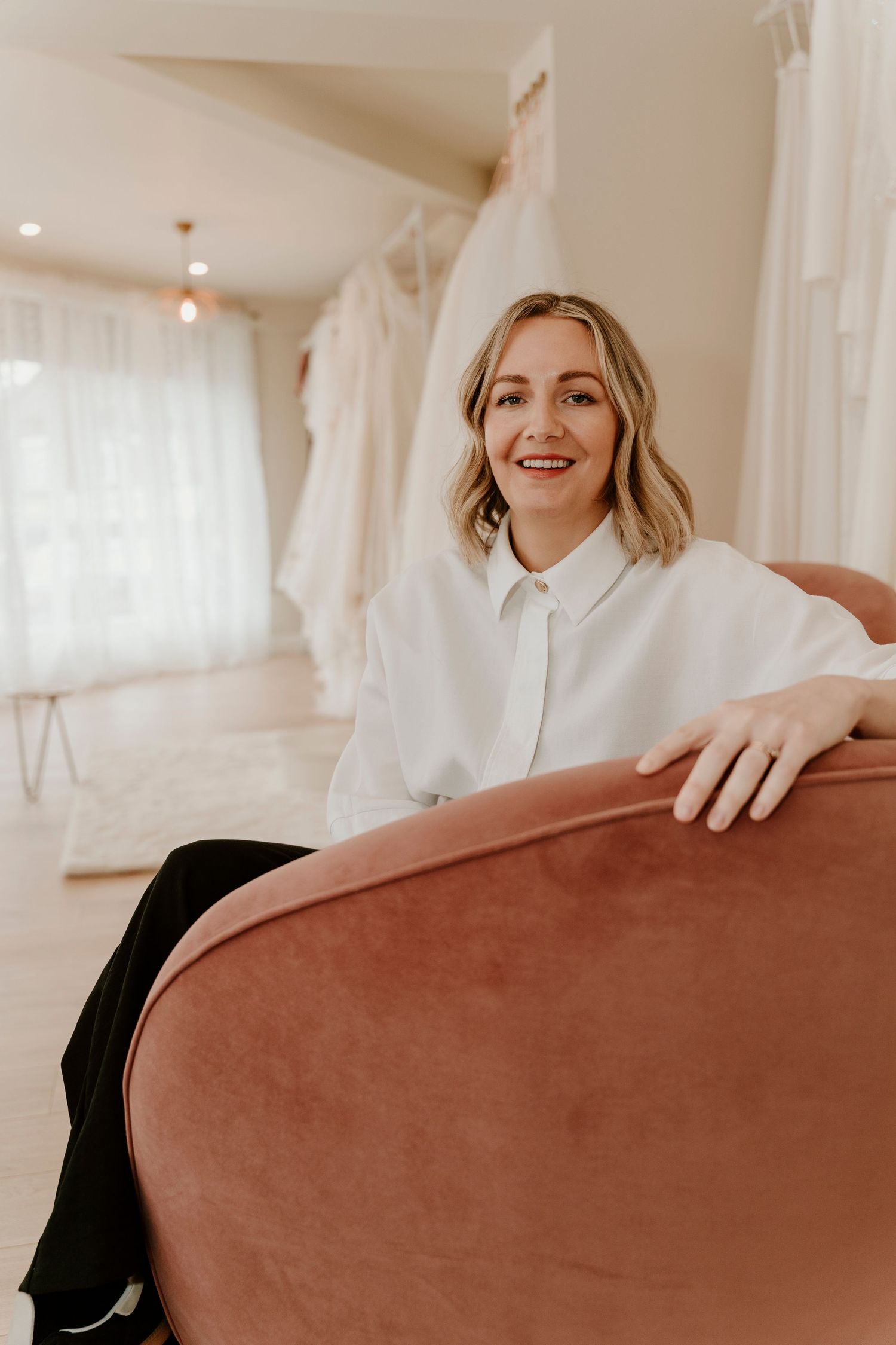 In a bridal boutique with white dresses in the background, a woman sits cheerfully on a pink velvet couch.