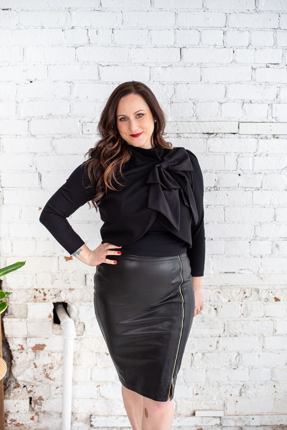 Fashionable black sweater with large bow detail paired with leather pencil skirt against white brick wall backdrop.