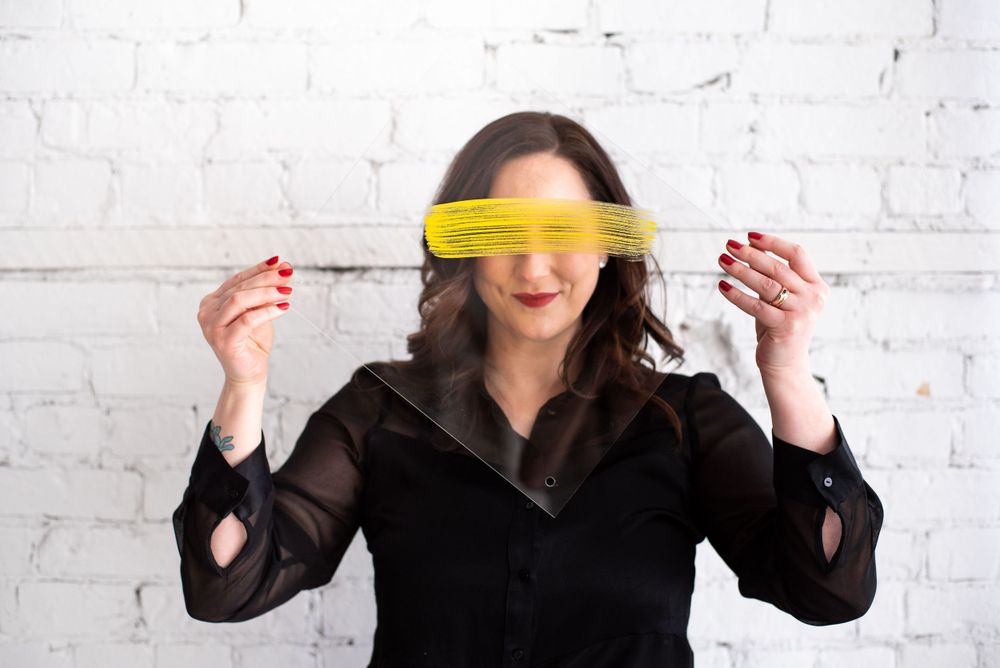 Person in black blouse holding yellow sticky note over eyes against white brick wall background.