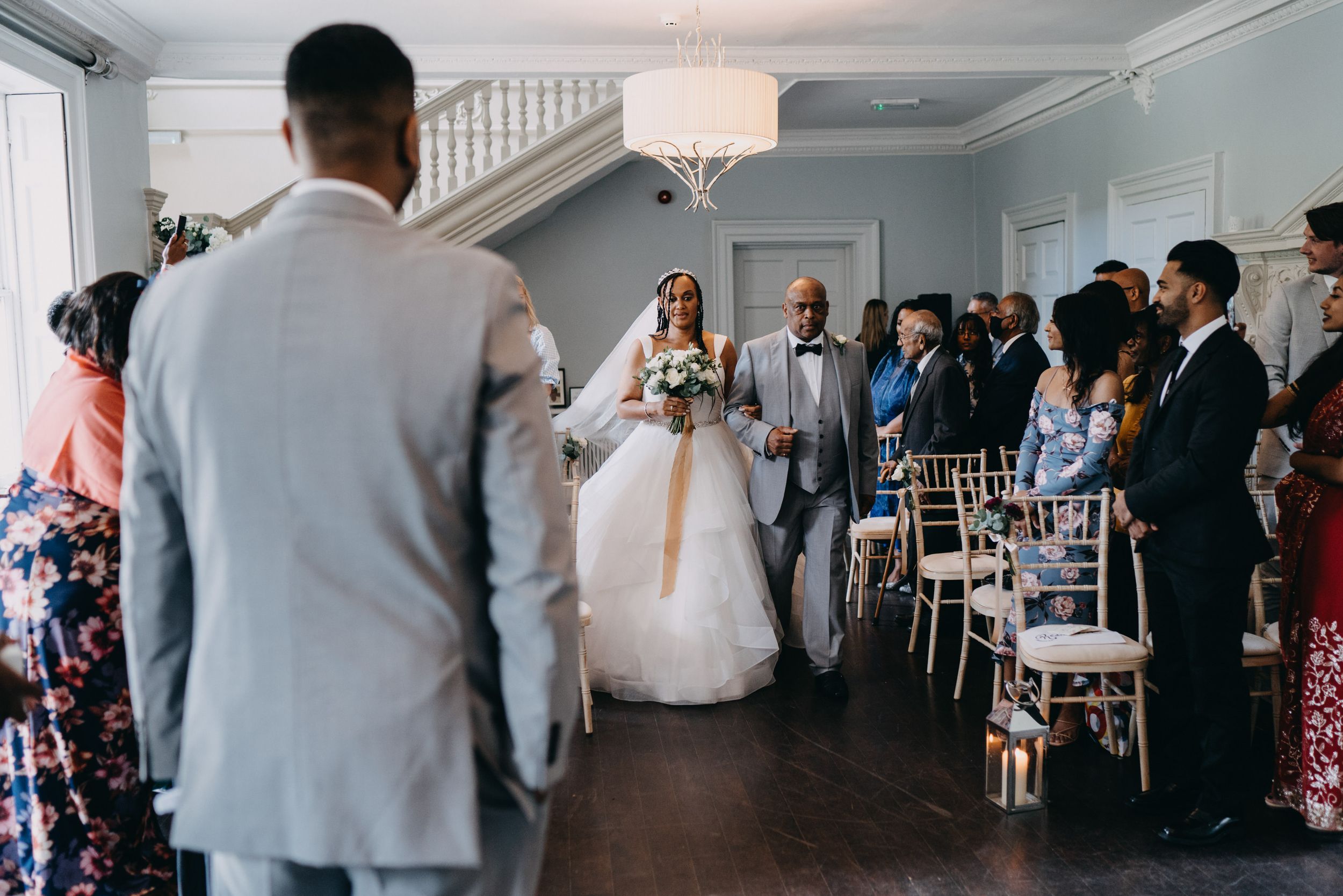 Wedding ceremony in a bright indoor venue with guests standing among white chairs as the bride walks down the aisle.