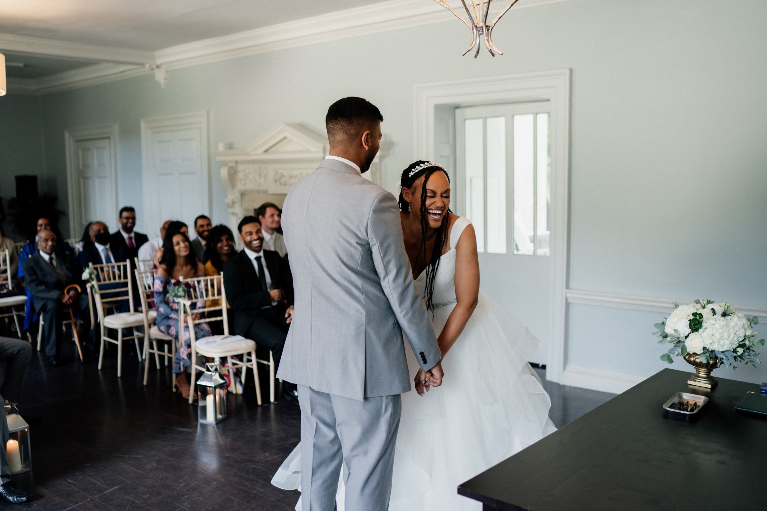 Two people stand together at the altar during an indoor wedding ceremony as guests look on in a bright white room.