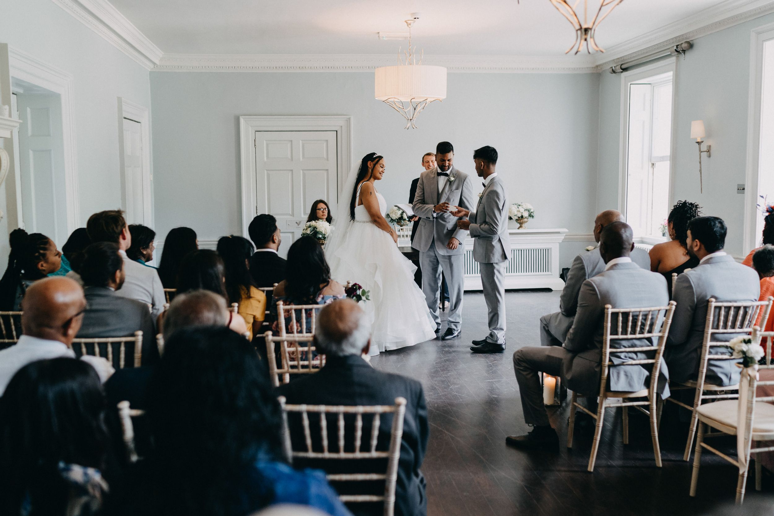 A small intimate wedding ceremony at Morden Hall , London takes place in a bright white room with chiavari chairs and guests looking on.