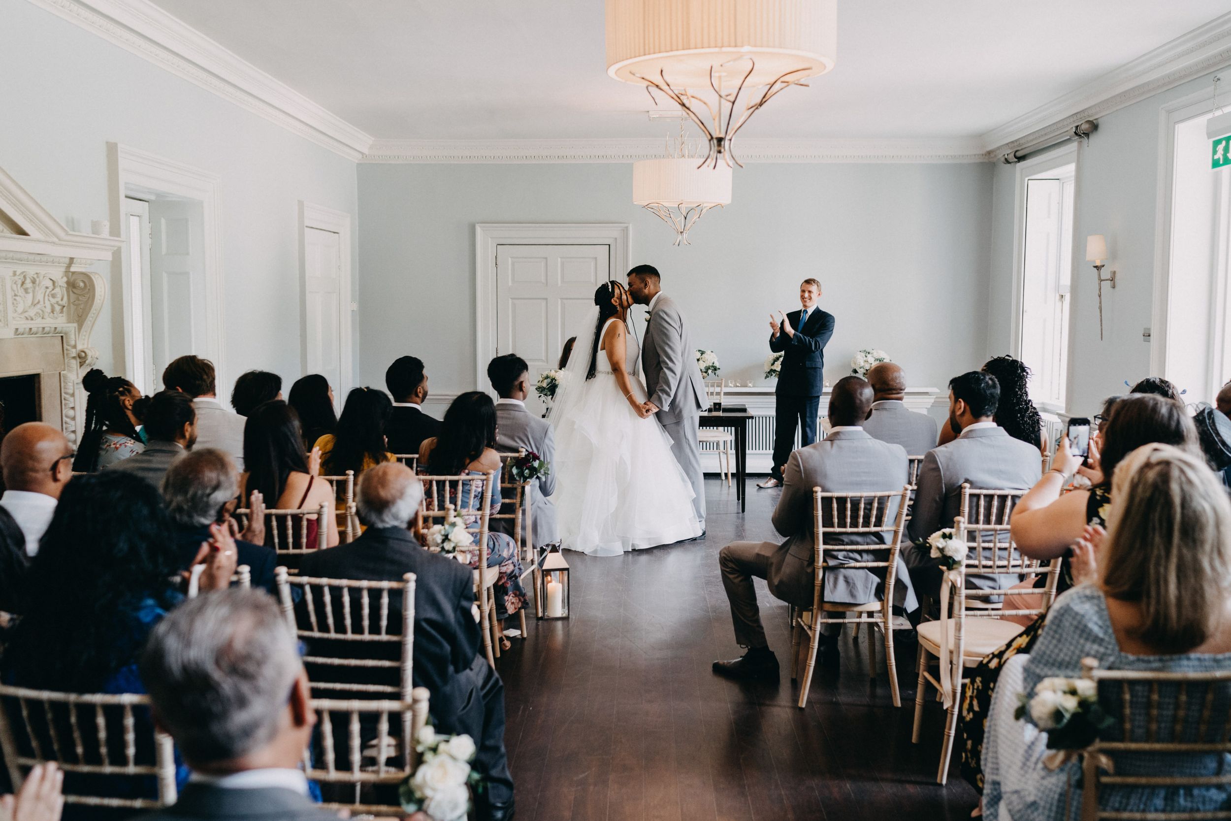 An intimate wedding ceremony takes place in a bright white room with guests seated in elegant Chiavari chairs.