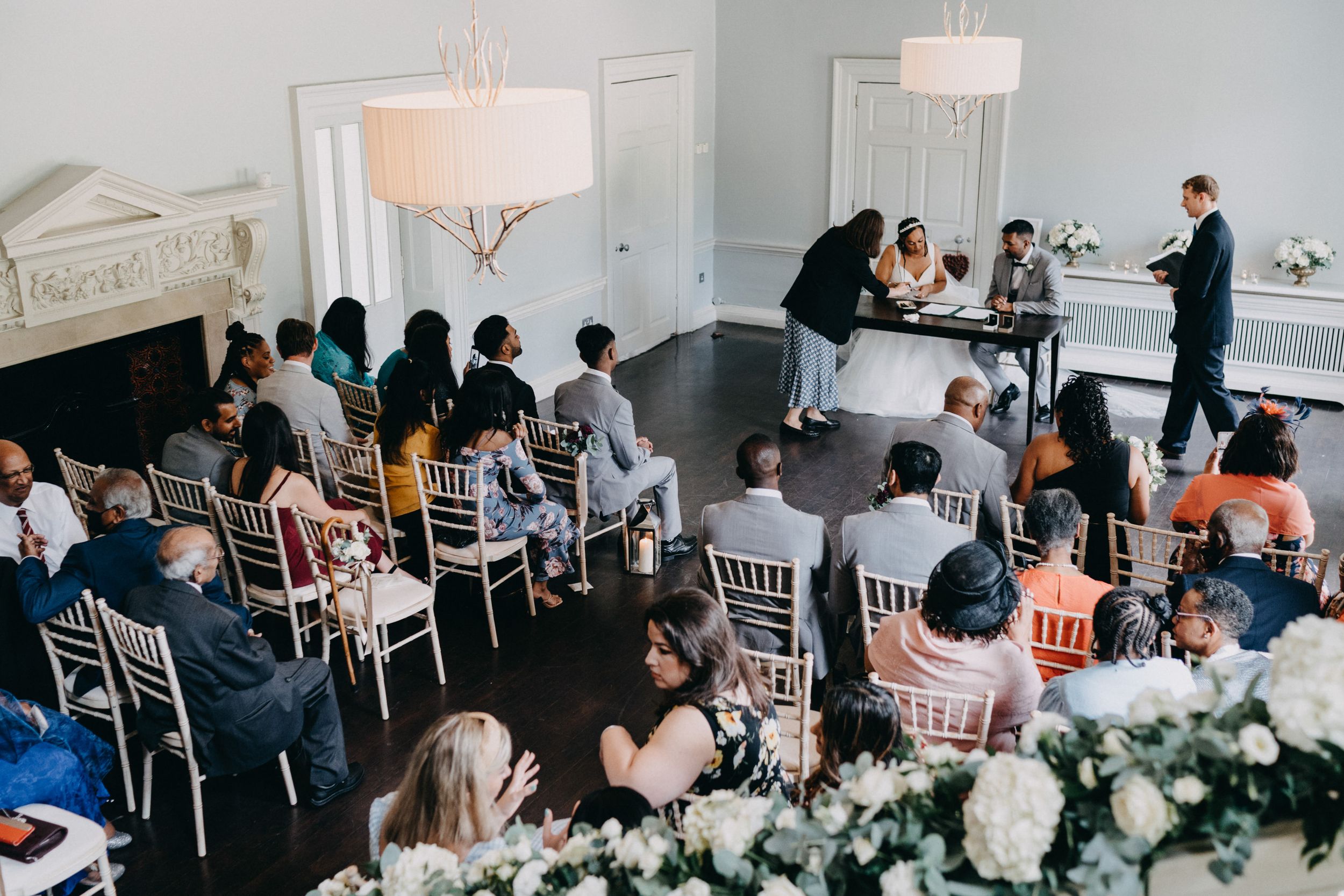 Wedding guests seated at elegant white chairs during an indoor reception at Morden Hall with floral arrangements and chandeliers.