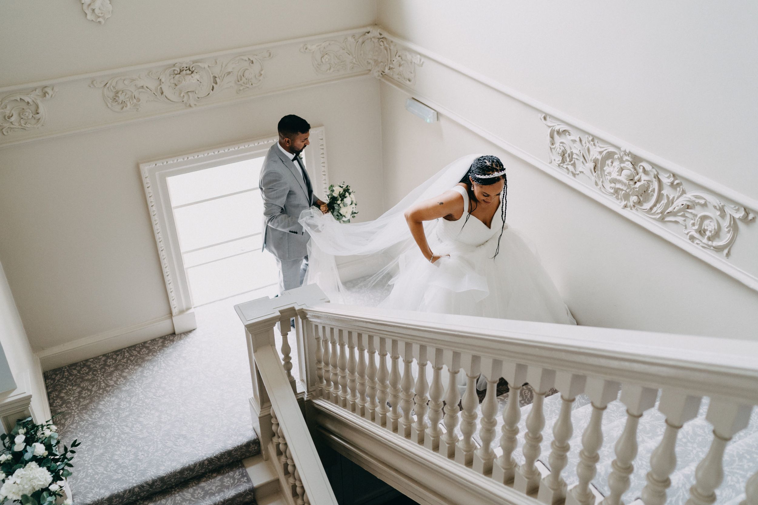 A couple in wedding attire poses on an elegant white staircase with ornate molding in a bright interior setting.