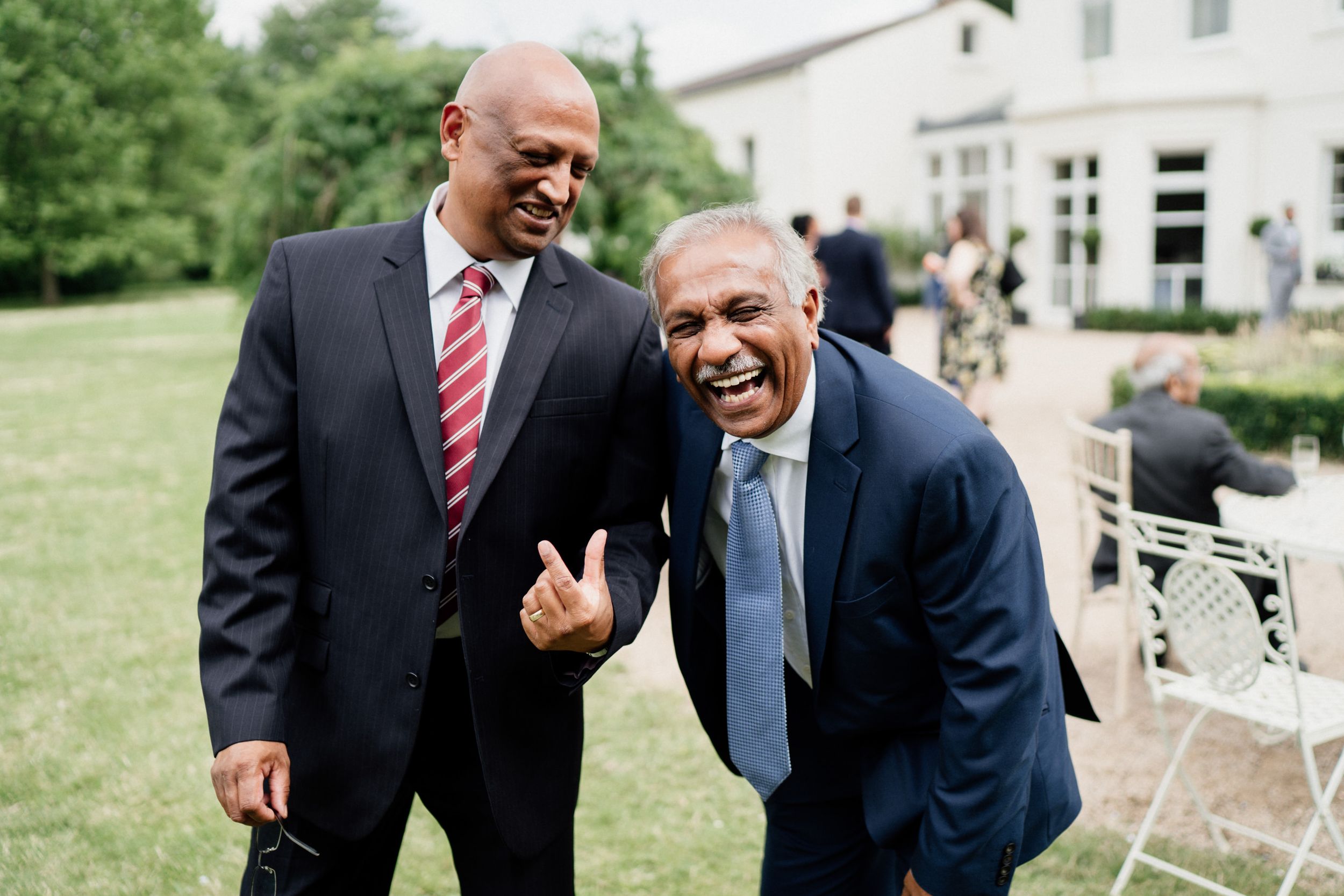 Two men in business suits share a lighthearted moment outdoors while laughing together on a sunny day.