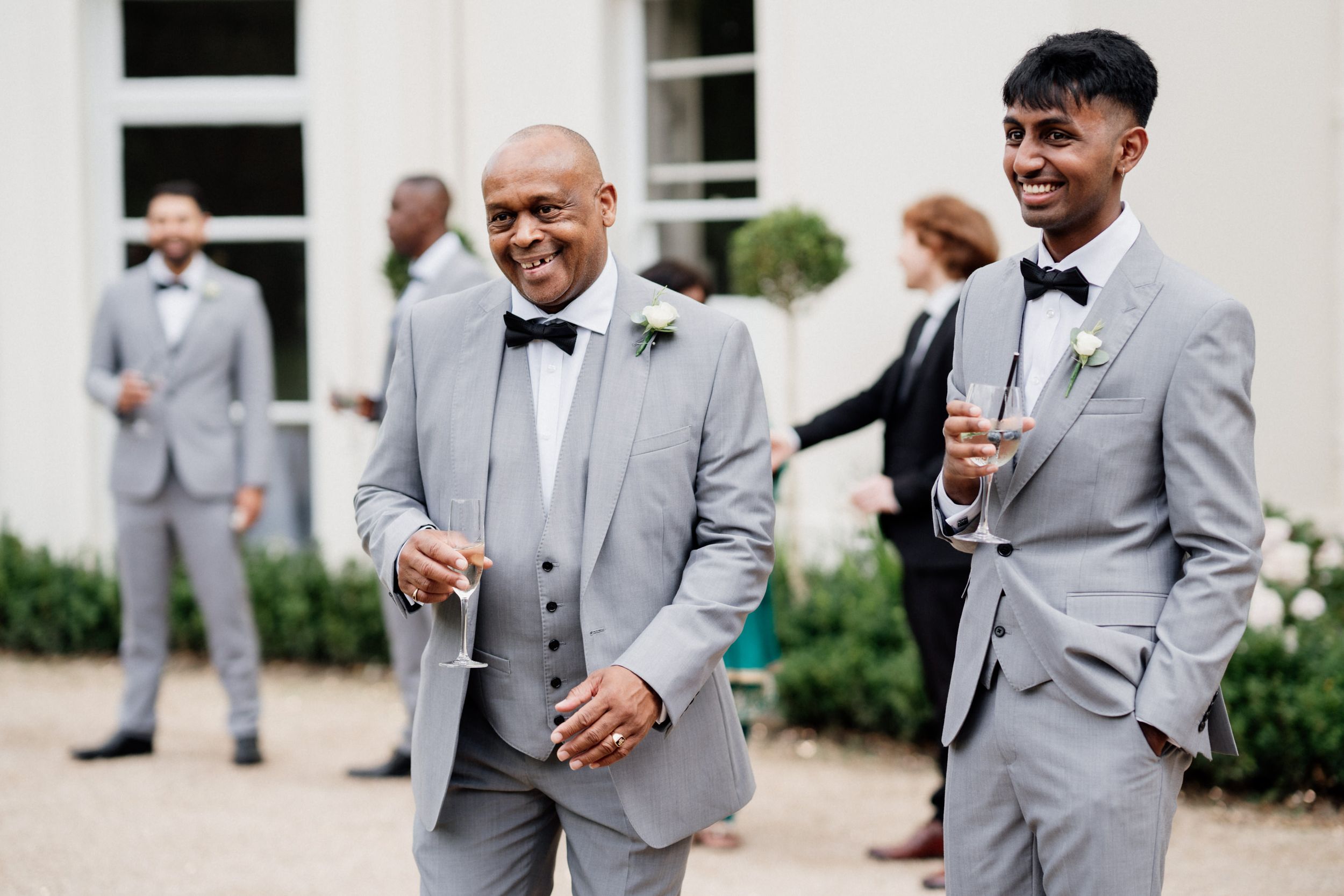 Two groomsmen share a laugh while dressed in gray suits and bow ties at an elegant outdoor wedding venue.
