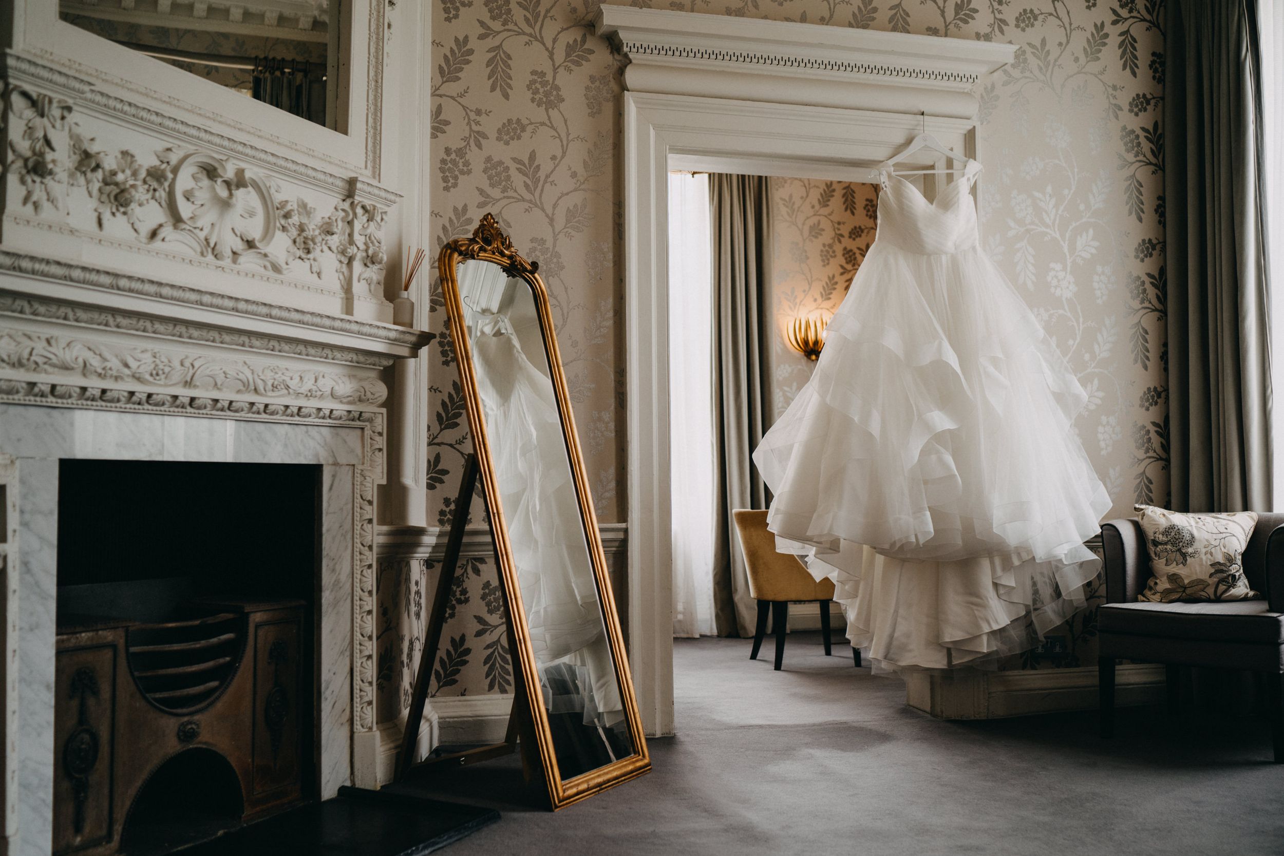 A white wedding dress hangs in an elegant room of Morden Hall with ornate moldings, vintage fireplace and gold-framed mirror.
