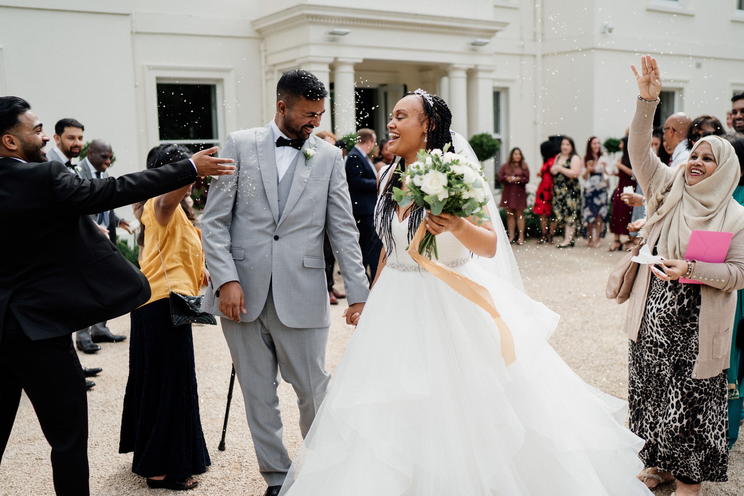 Joyful wedding celebration outside white building with couple walking while guests cheer and throw confetti.