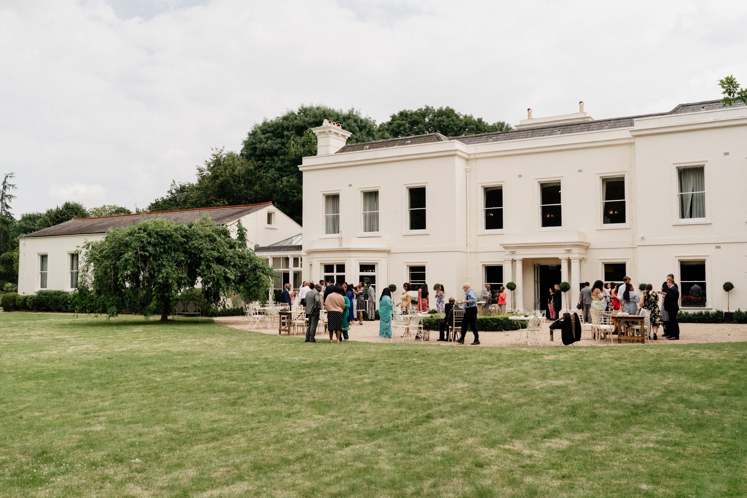 Guests gather outside Morden Hall an elegant white Georgian-style building with large windows on a grassy lawn during an outdoor event.