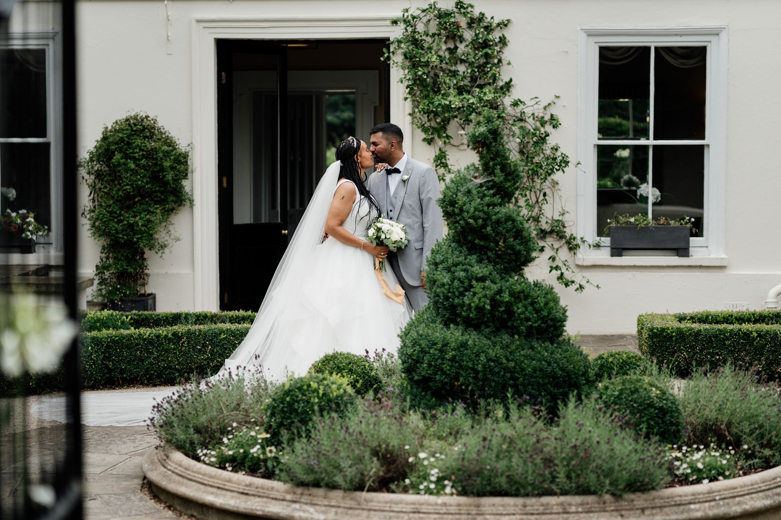 A couple in wedding attire sharing a moment in a garden with manicured hedges and climbing vines against a white building.