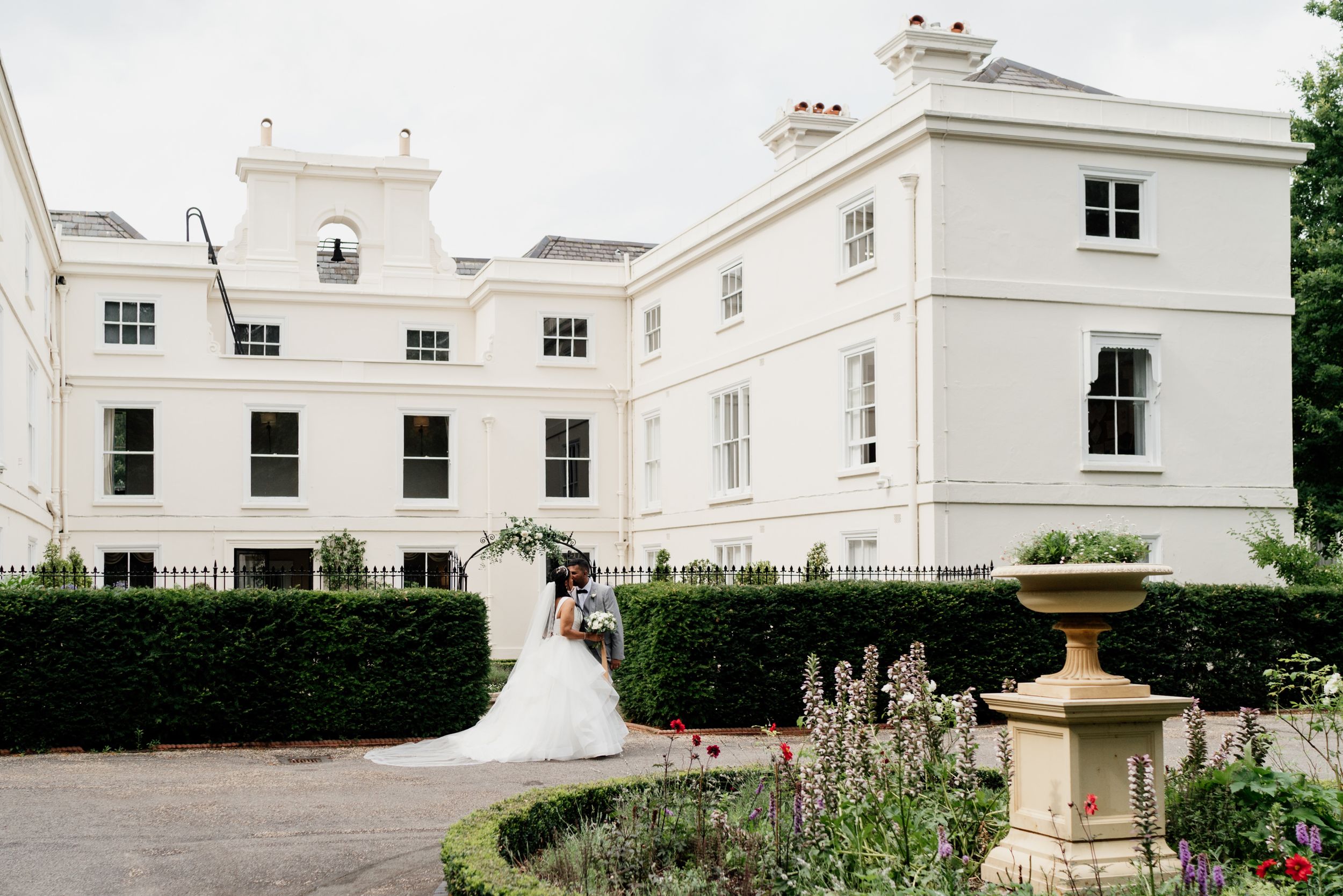 A grand white Georgian manor house of Morden Hall in London with manicured gardens and stone fountain in the foreground.