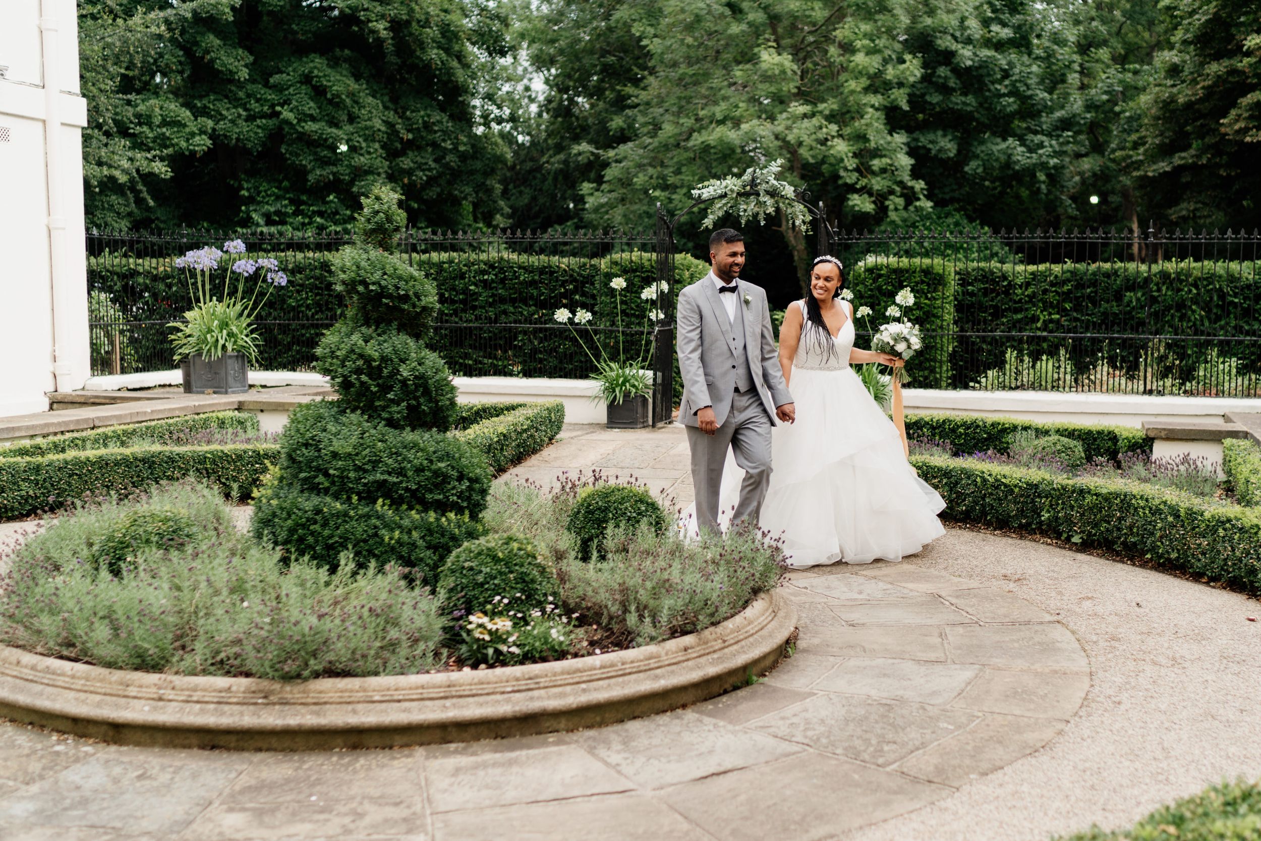 Wedding couple standing together in an elegant formal garden of London Morden Hall with manicured shrubs and curved stone pathways.