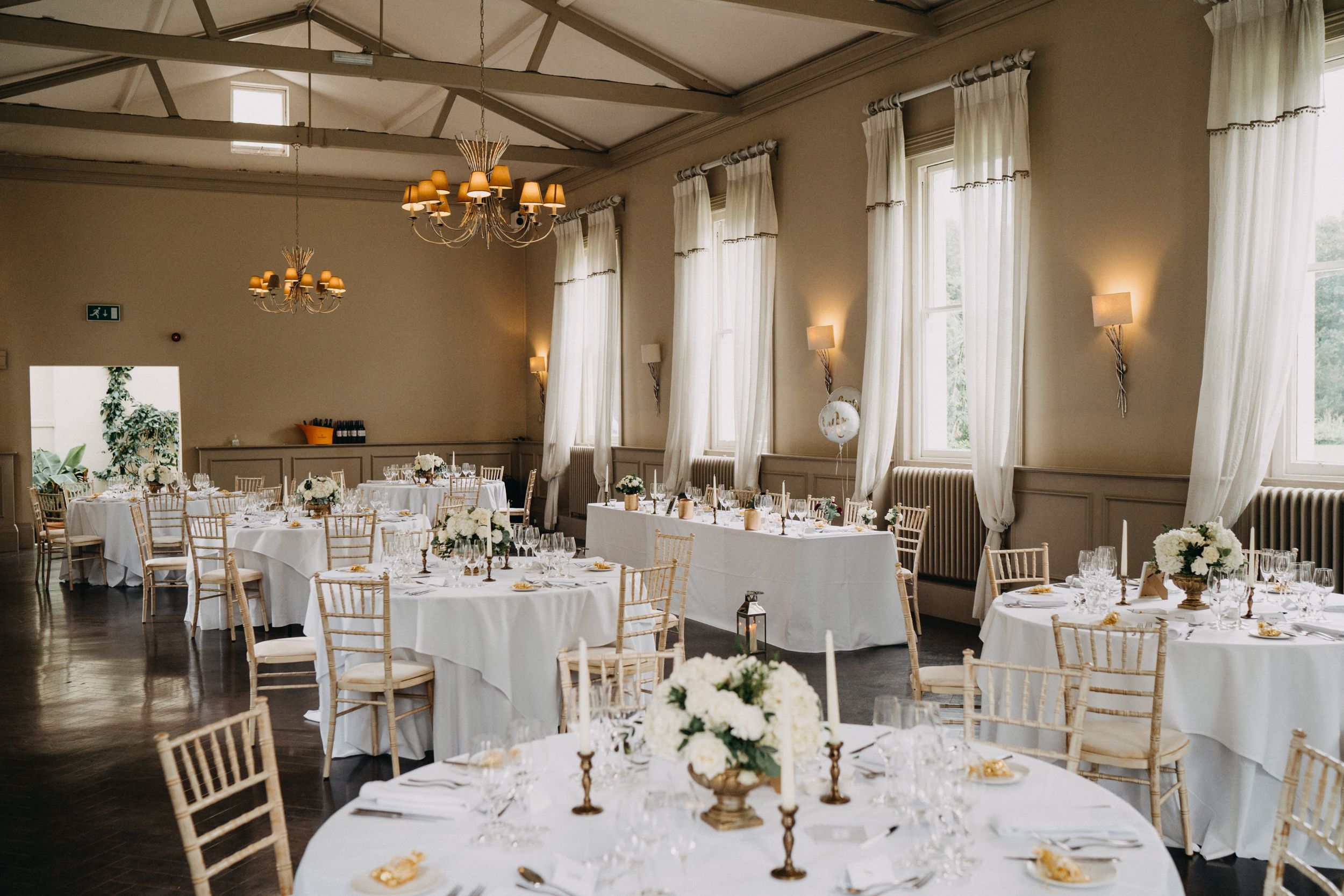 Elegant wedding reception hall at Morden Hall with white tablecloths, chiavari chairs, and crystal chandeliers in natural light.
