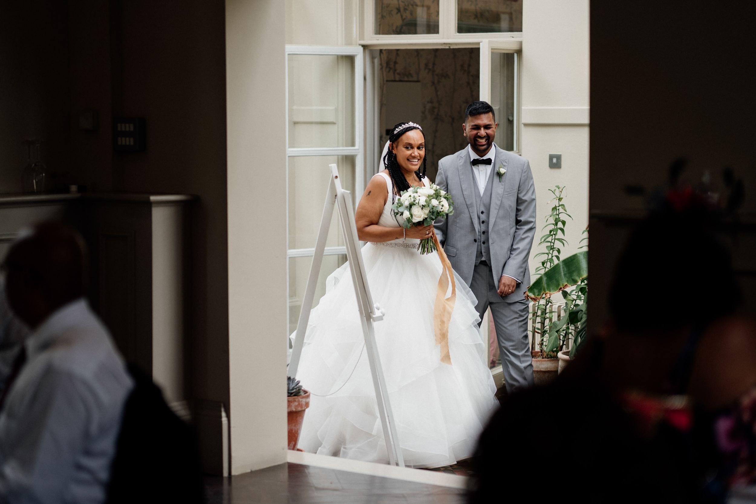 Two wedding figures stand in a white doorway on their special day, with the bride in a flowing white gown.
