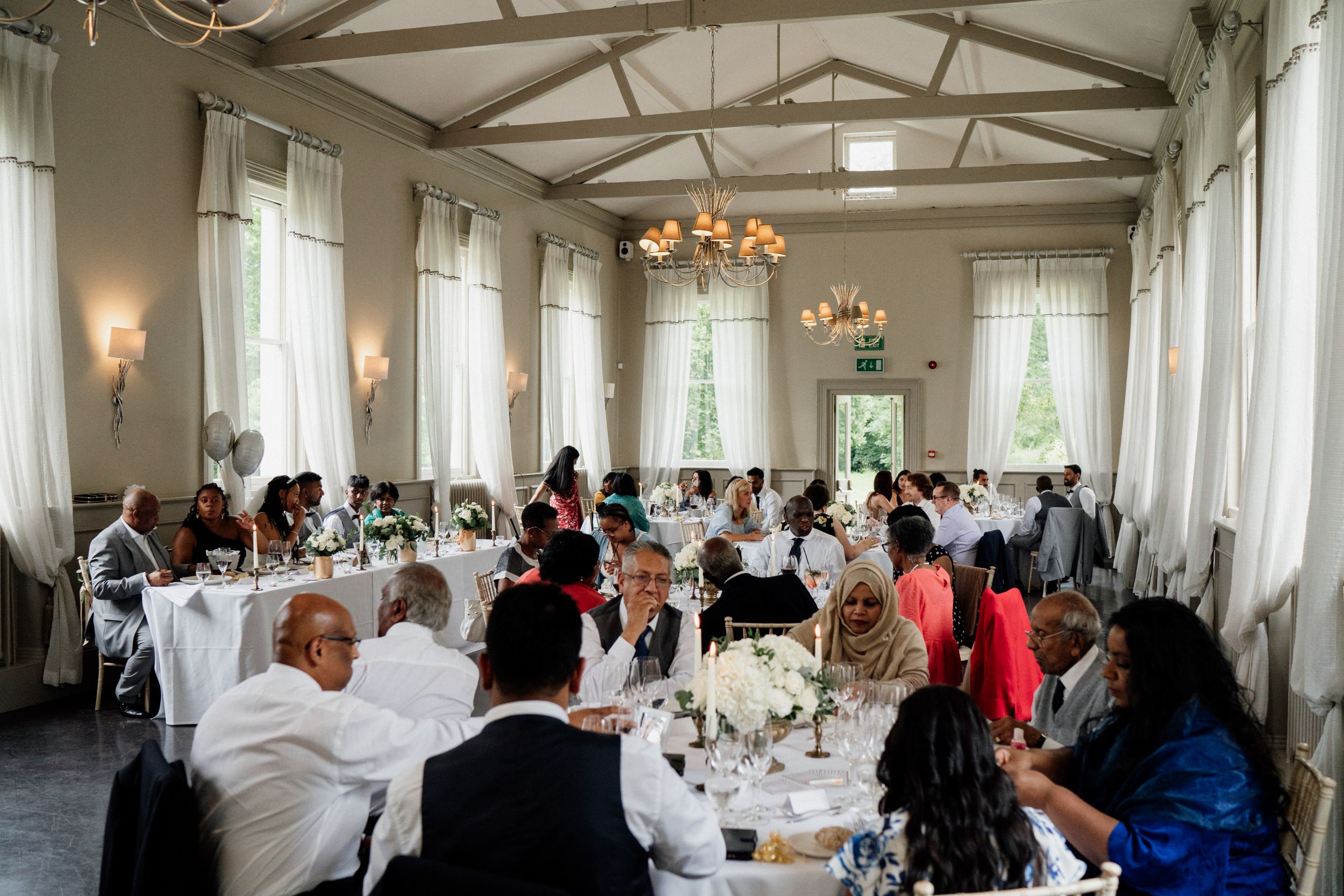 Wedding reception in an elegant white room with exposed wooden beams and large windows where guests dine at round tables.