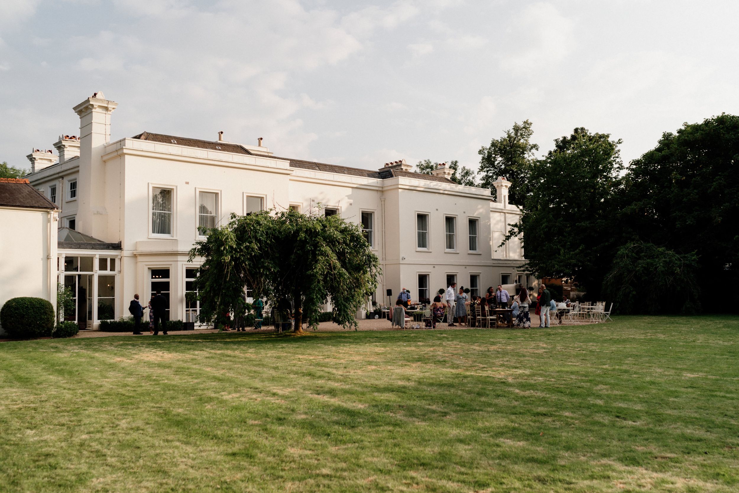 Elegant white manor house of Morden Hall with guests gathered on the lawn for an outdoor event on a summer day.