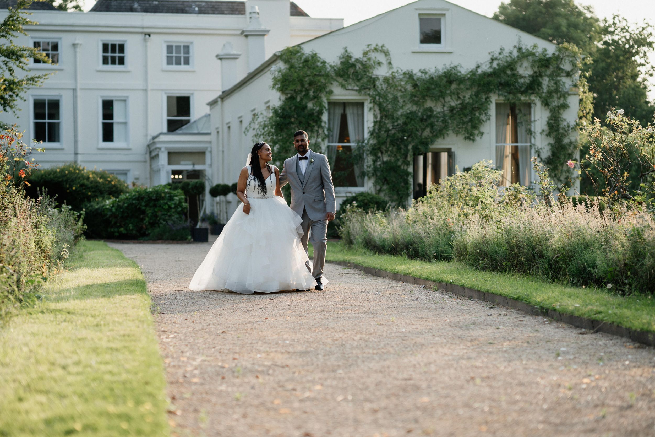 Newlyweds walk together on a gravel path beside a white mansion and garden during golden hour.