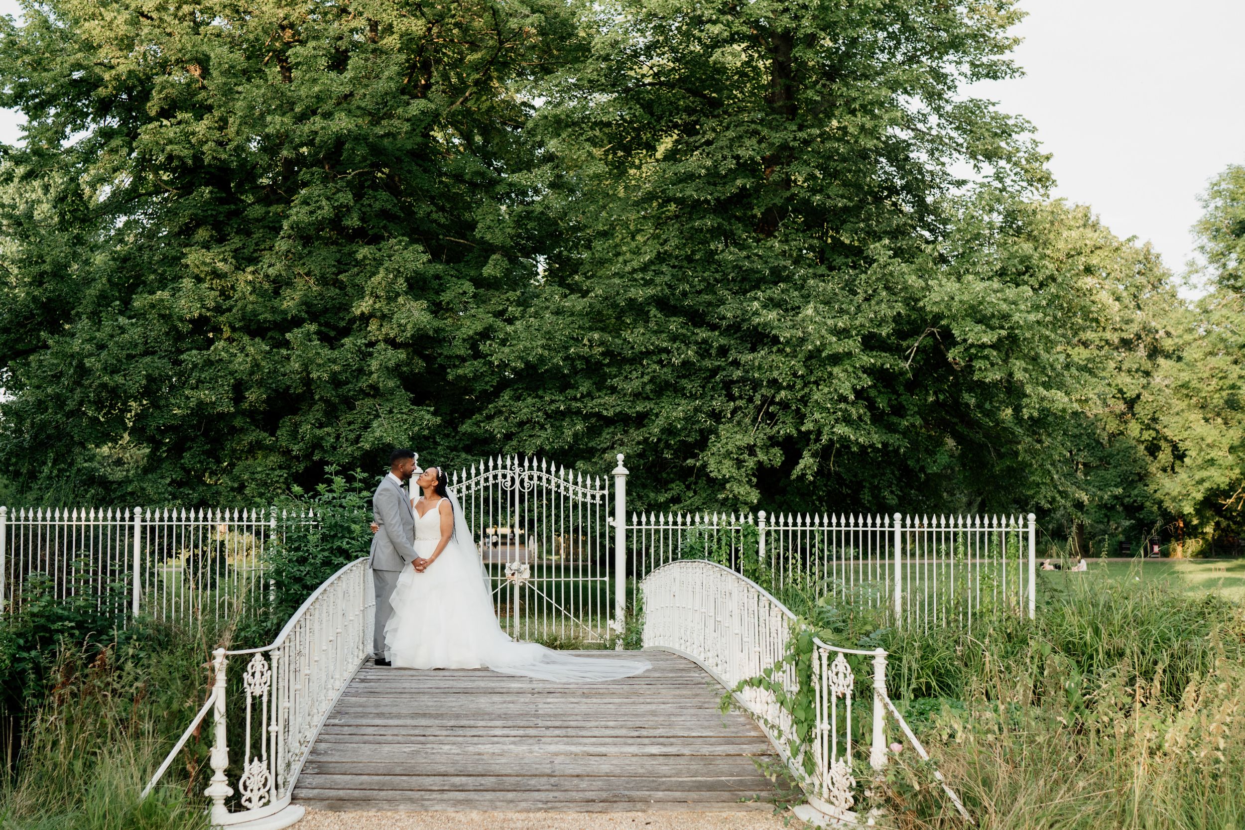 Couple standing on a white wooden footbridge surrounded by lush greenery in a garden setting of Morden Hall Gardens.