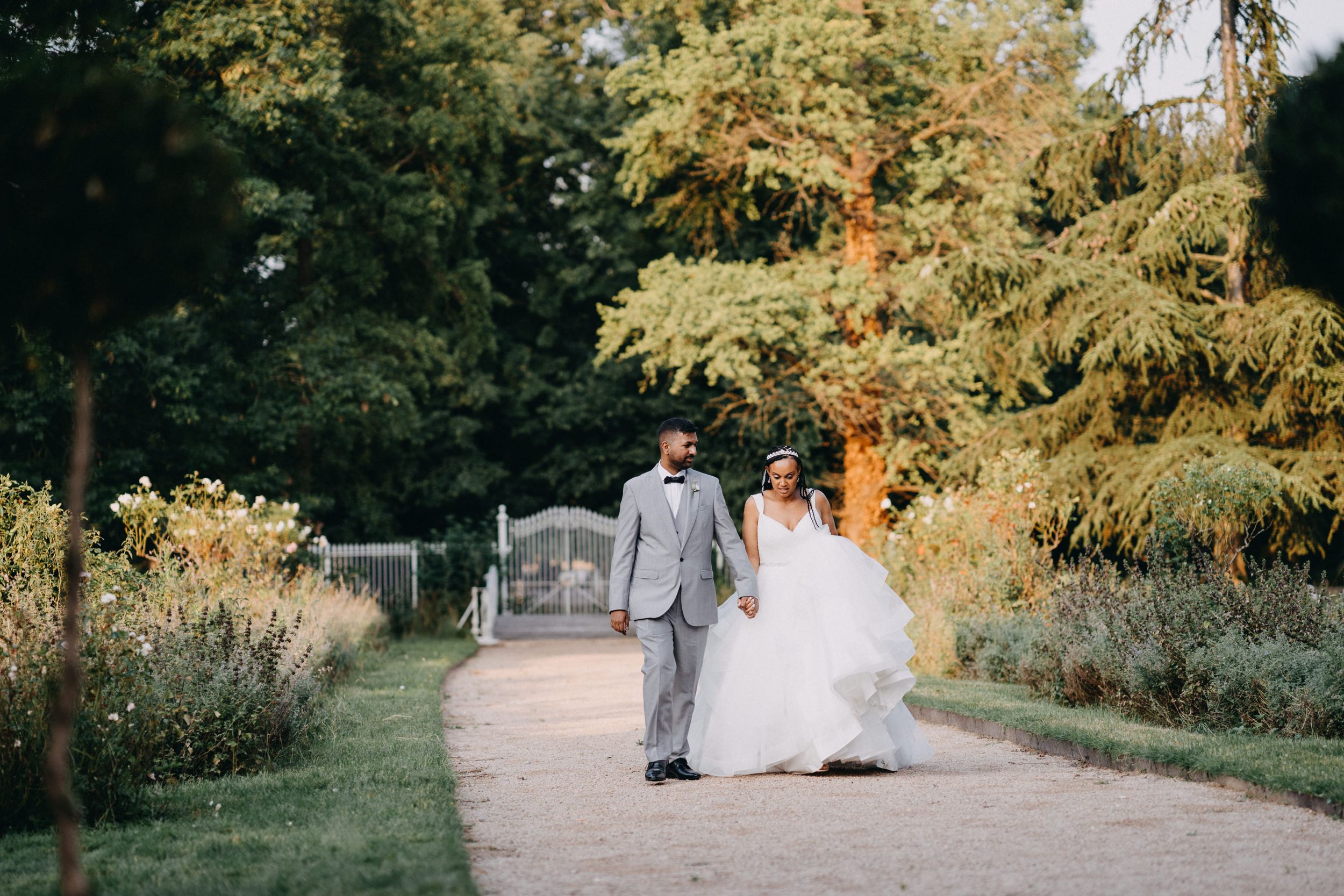 A couple walks down a garden path surrounded by lush autumn foliage on a sunny day.