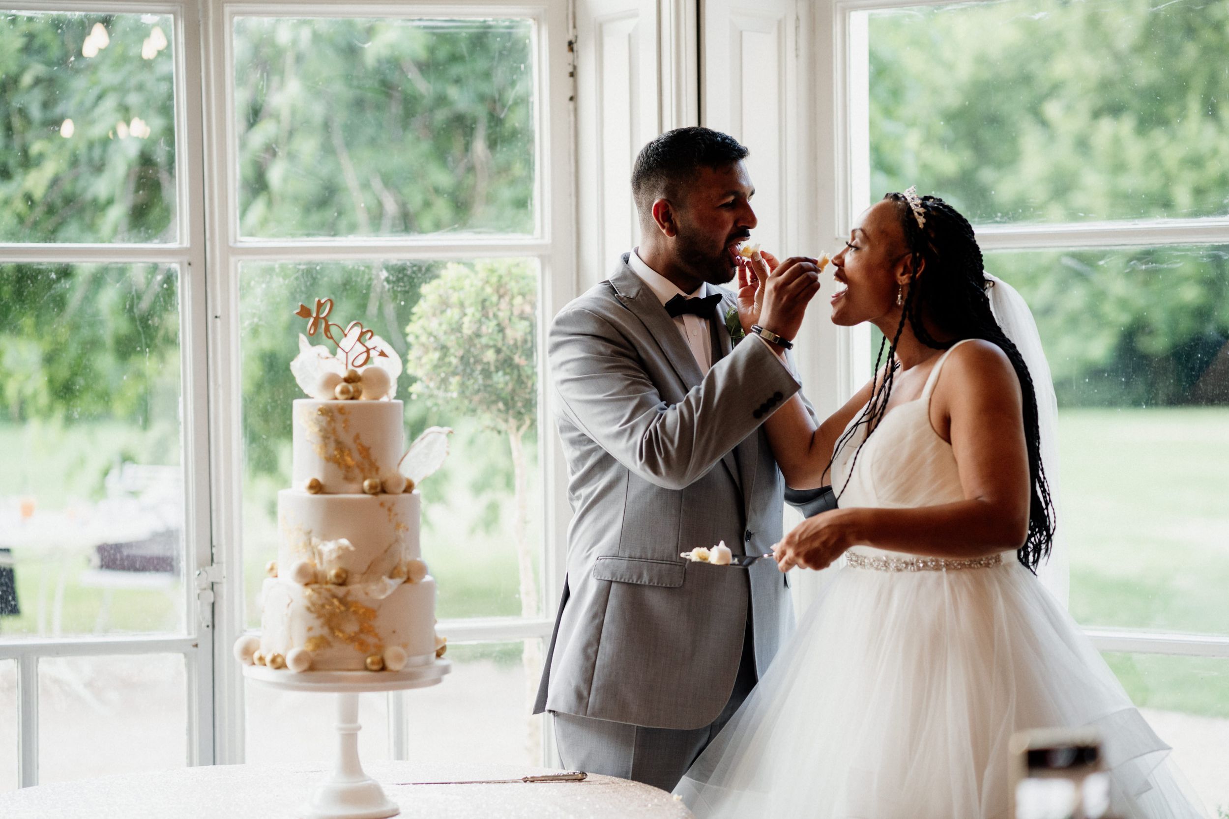 A newlywed couple feeds each other wedding cake during their reception in a bright sunlit room with a floral cake display.