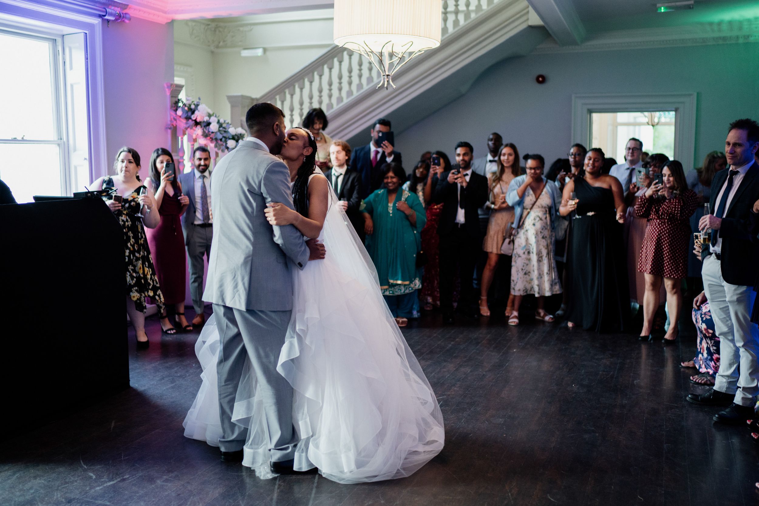 Couple shares their first dance as newlyweds while guests watch at an indoor wedding reception with purple lighting.