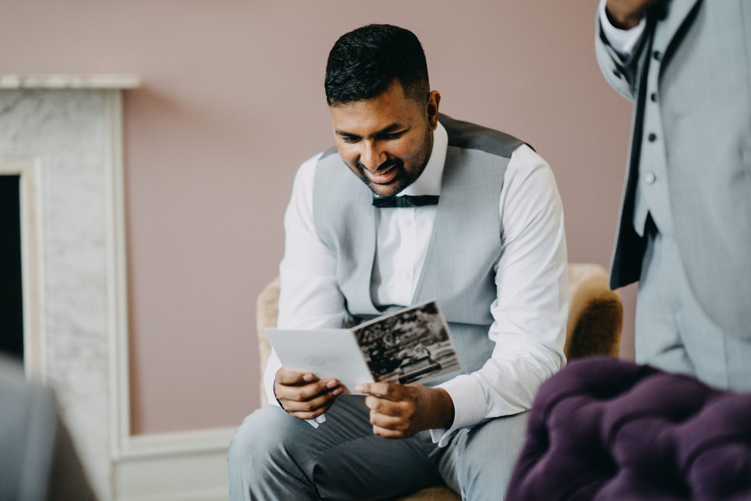 A person in formal attire looking at a vintage camera while sitting on a couch in a pink-walled room.