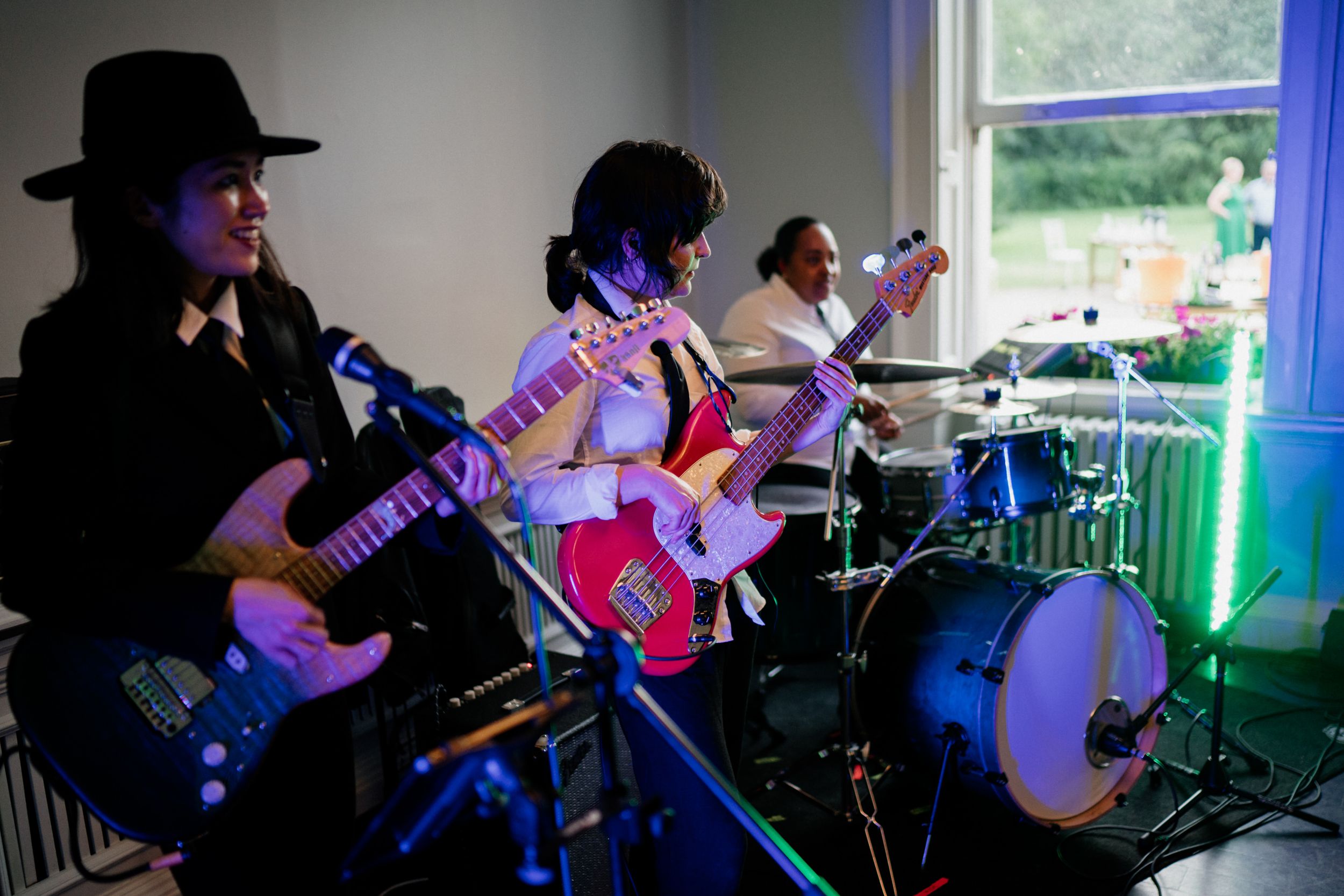 A live band performs on stage with guitarists in dark attire and a drummer playing in colorful lighting.